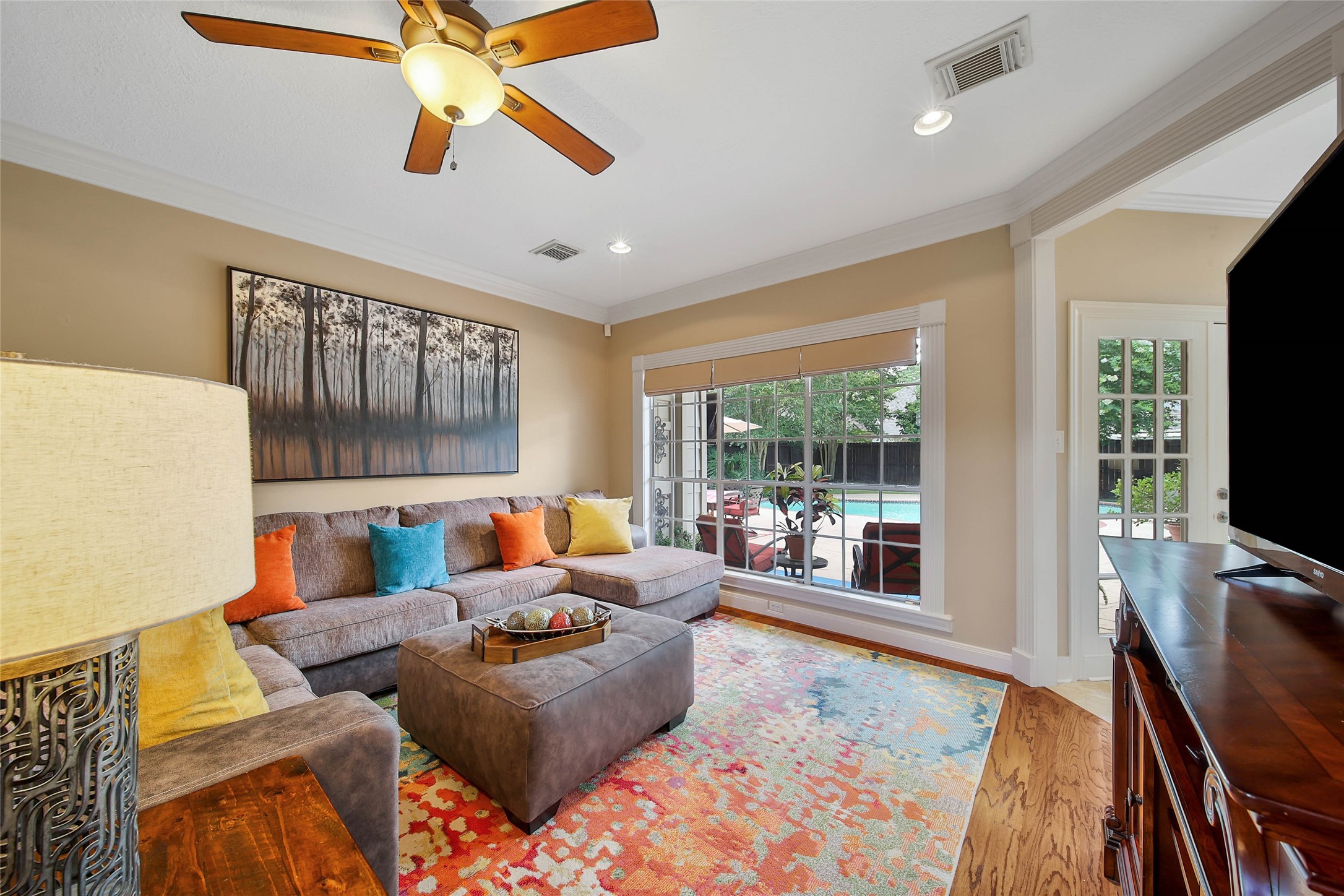 This inviting living room features a cozy sectional sofa with colorful cushions, a vibrant area rug, and a ceiling fan for comfort. Large windows offer a view of the backyard pool area, bringing in natural light. The space is enhanced by hardwood floors and tasteful wall art.