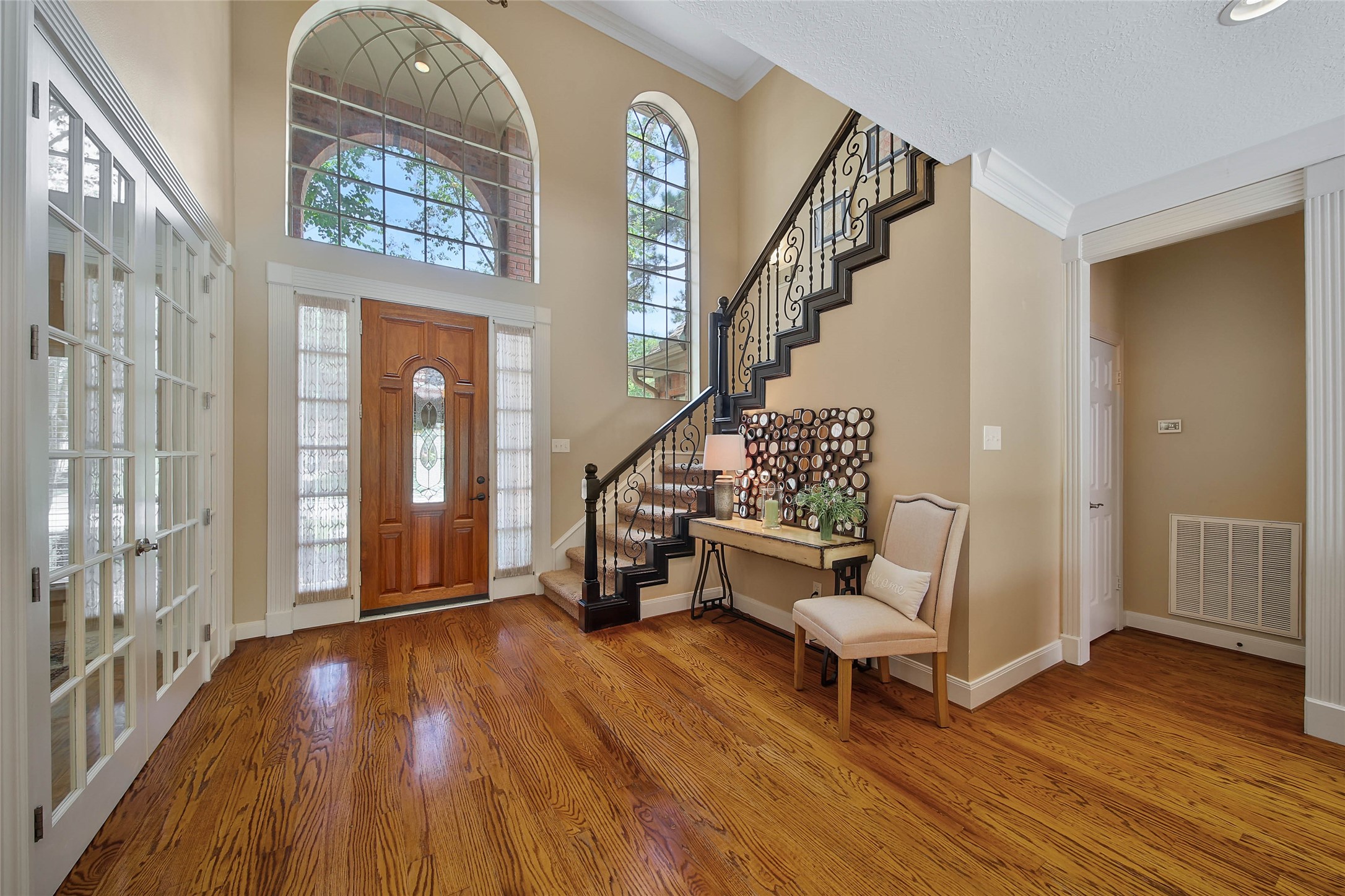 Spacious entryway with elegant wood flooring and a grand staircase. Large, arched windows provide ample natural light, and French doors lead to an adjacent room. The space is warm and inviting, perfect for welcoming guests.
