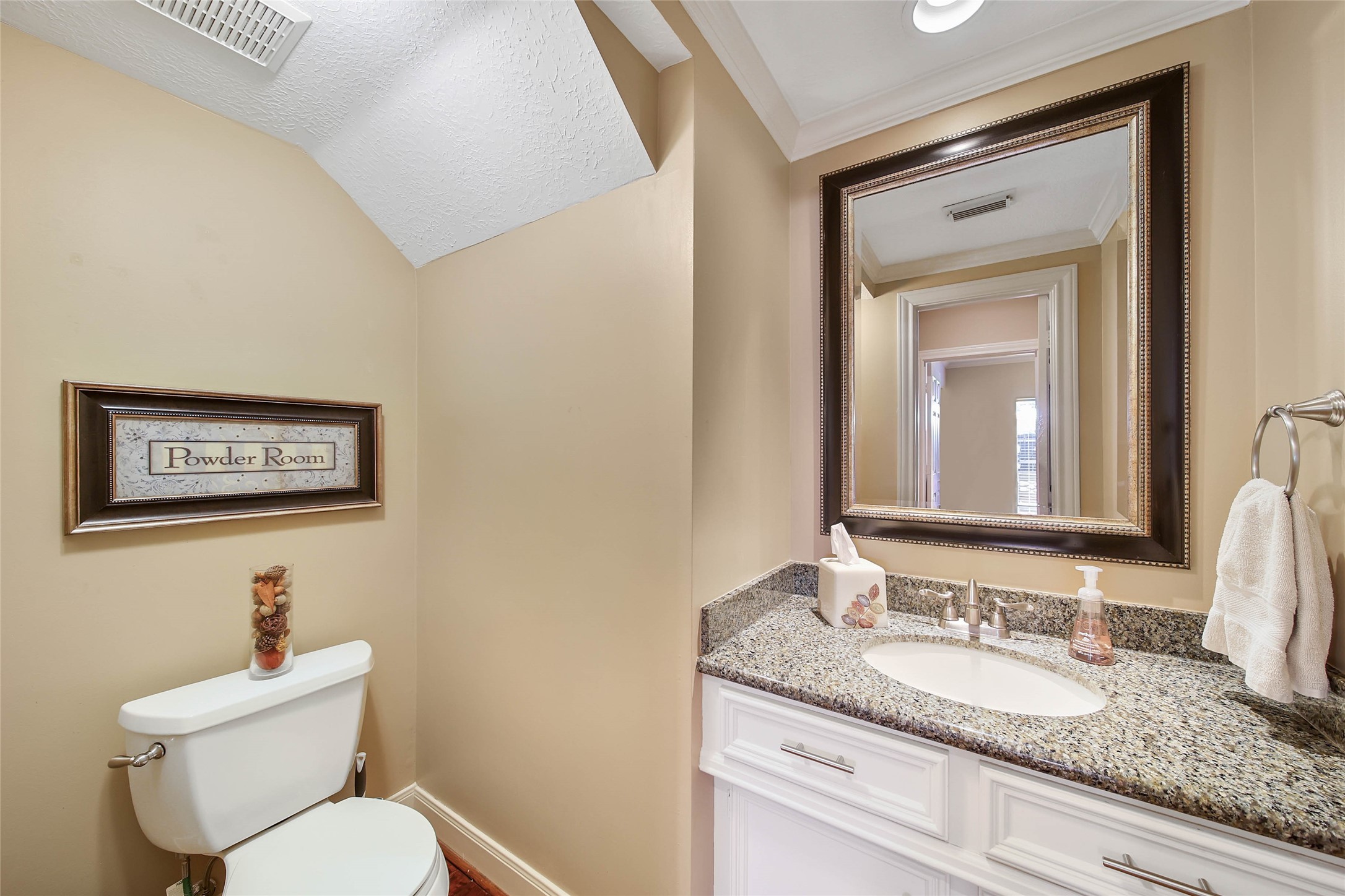 This powder room features a sleek granite countertop, a large framed mirror, and warm beige walls. It includes a toilet with decorative accents and a towel ring, creating a cozy and inviting space.