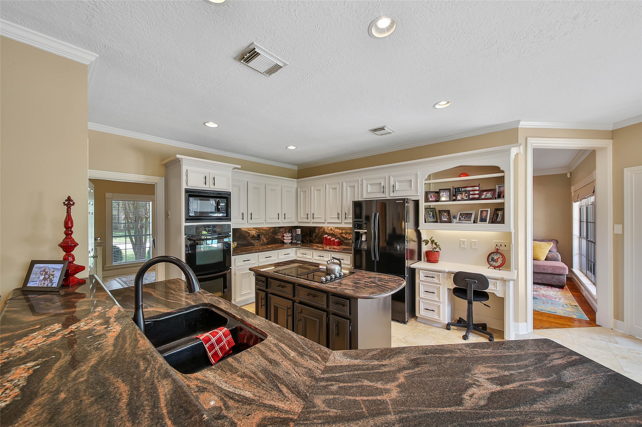 Spacious kitchen with elegant granite countertops, a central island, and modern black appliances. It features white cabinetry, a built-in desk area, and opens to a cozy living space. Ideal for both cooking and entertaining.