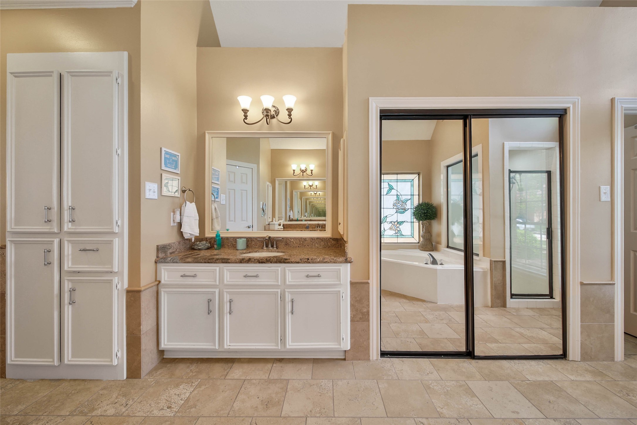 This photo showcases a spacious, elegant bathroom with neutral tones. It features a double vanity with ample storage, a large mirror, and stylish lighting. A glass door leads to a serene bathtub area with a decorative window, creating a luxurious feel.