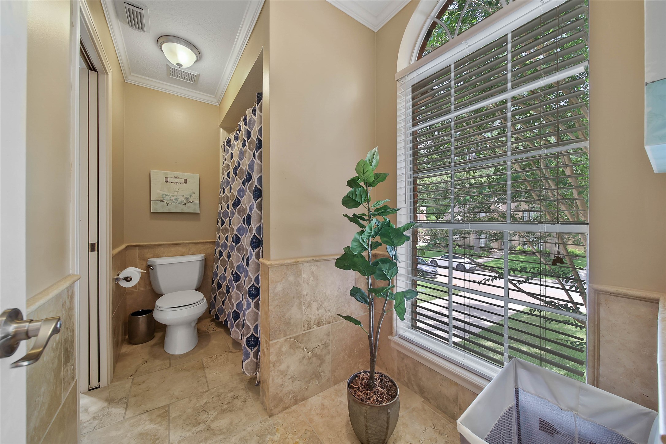 This bathroom features a tiled floor, a toilet, and a shower with a patterned curtain. A large window with blinds provides natural light, and there's a decorative plant for a fresh touch.