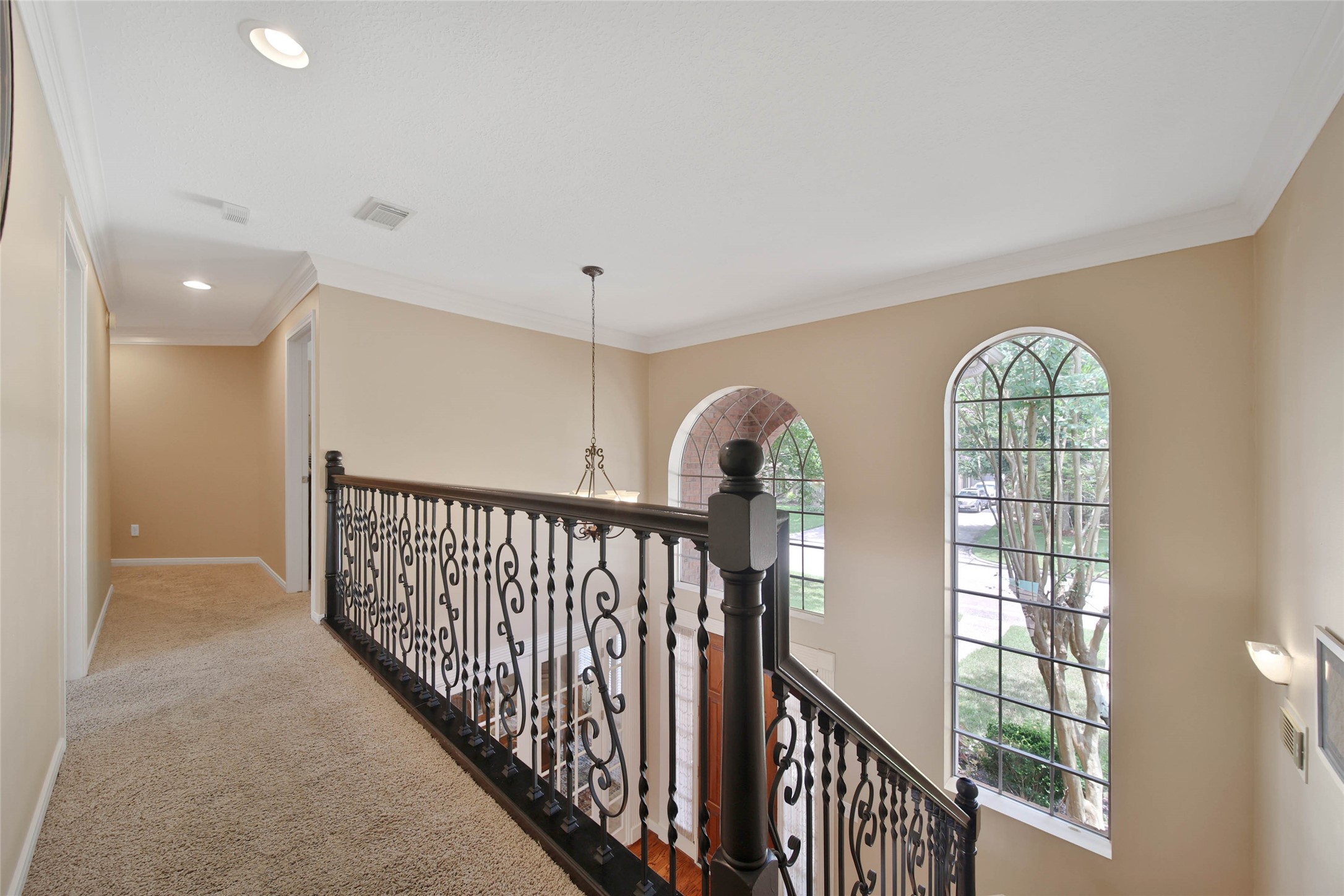 This photo shows an elegant upstairs hallway with a wrought-iron railing overlooking large arched windows that provide natural light. The space is carpeted and features soft, neutral walls, creating a warm and inviting atmosphere.