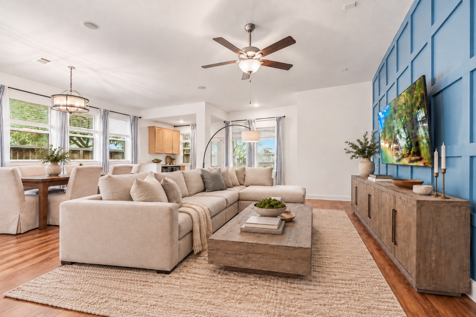 Spacious living area with elegant wood flooring, featuring a stylish accent wall. Abundant natural light streams through large windows. Virtually Staged