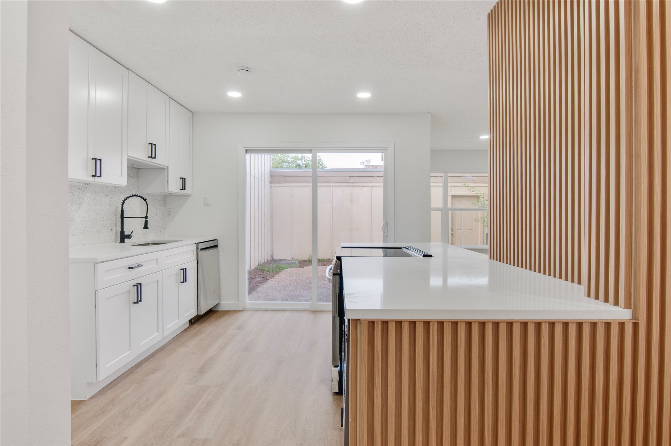 Modern kitchen with sleek white cabinetry, stylish black fixtures, and a spacious island. Natural light pours in through sliding glass doors, highlighting the warm wood flooring and unique vertical slat design.