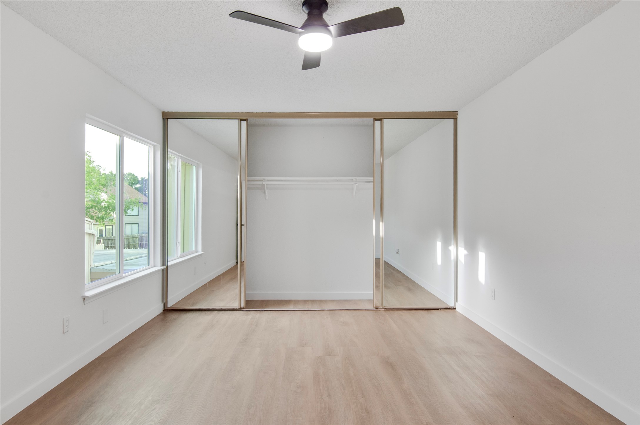 Bright bedroom featuring sleek mirrored closet doors, modern ceiling fan, and abundant natural light from large windows.
