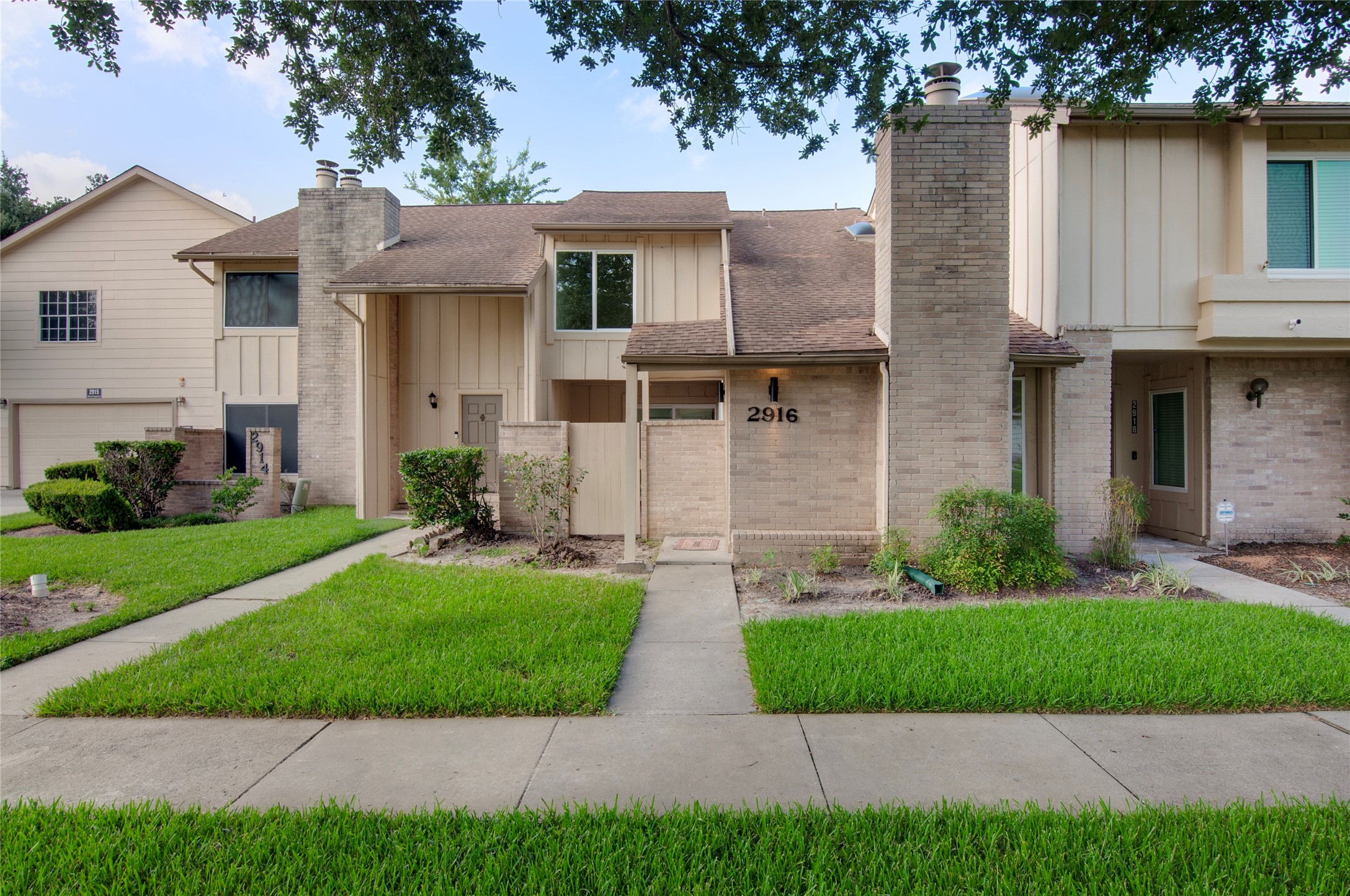 Charming two-story townhouse with a brick facade and a welcoming front entrance, surrounded by lush greenery and shaded by mature trees.
