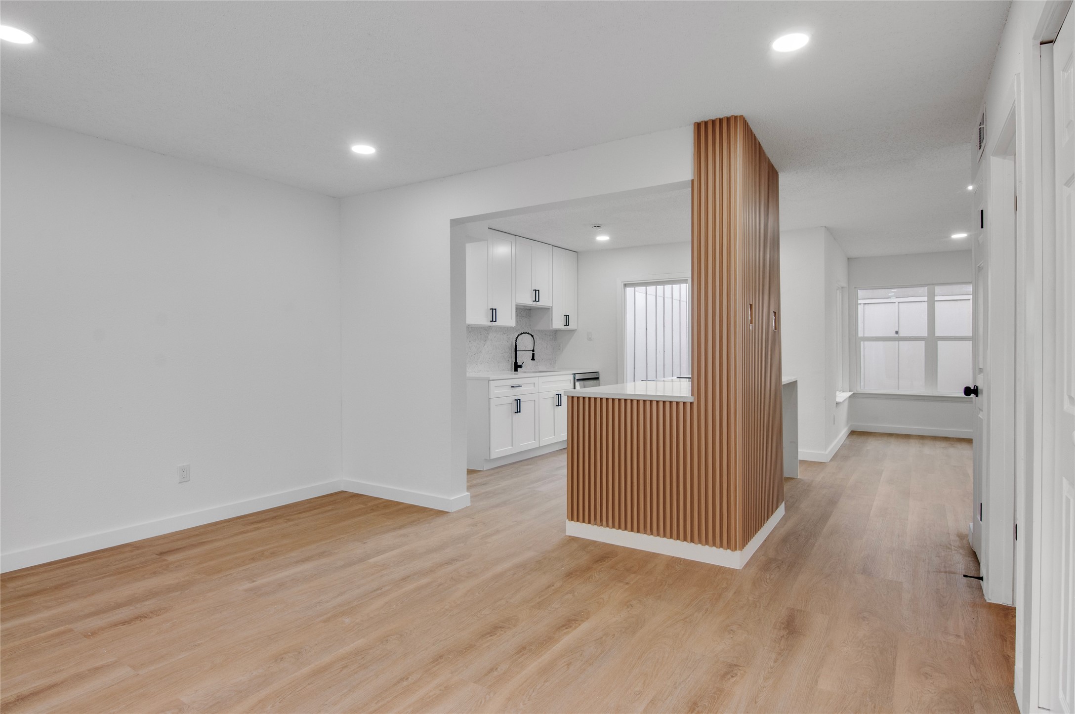Step into a modern open-concept living space featuring sleek wood flooring, a stylish accent wall, and a bright, contemporary kitchen with white cabinetry.