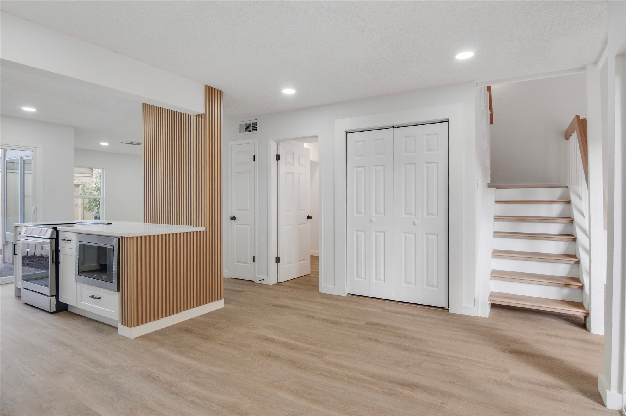 Modern open-concept space featuring sleek wood flooring, a stylish partition, and bright white walls. The area includes a contemporary kitchen with new stainless steel appliances and a convenient staircase leading to the upper level.