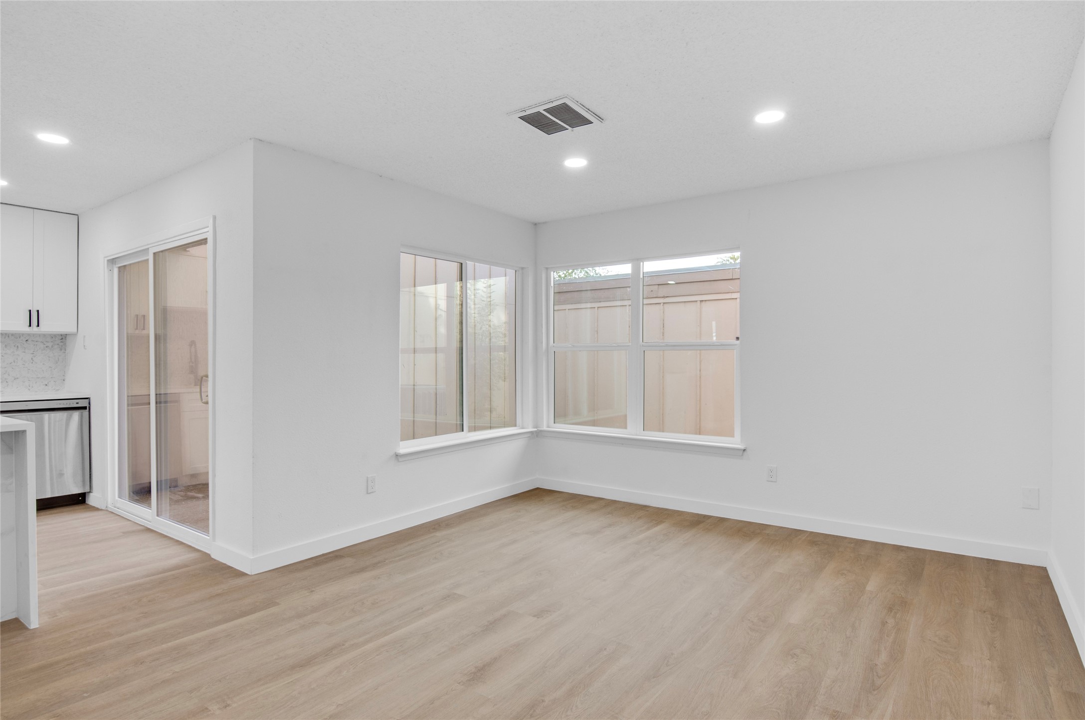 Bright and inviting living area with sleek wood flooring, recessed lighting, and large windows offering ample natural light, seamlessly connecting to a modern kitchen with stainless steel appliances.