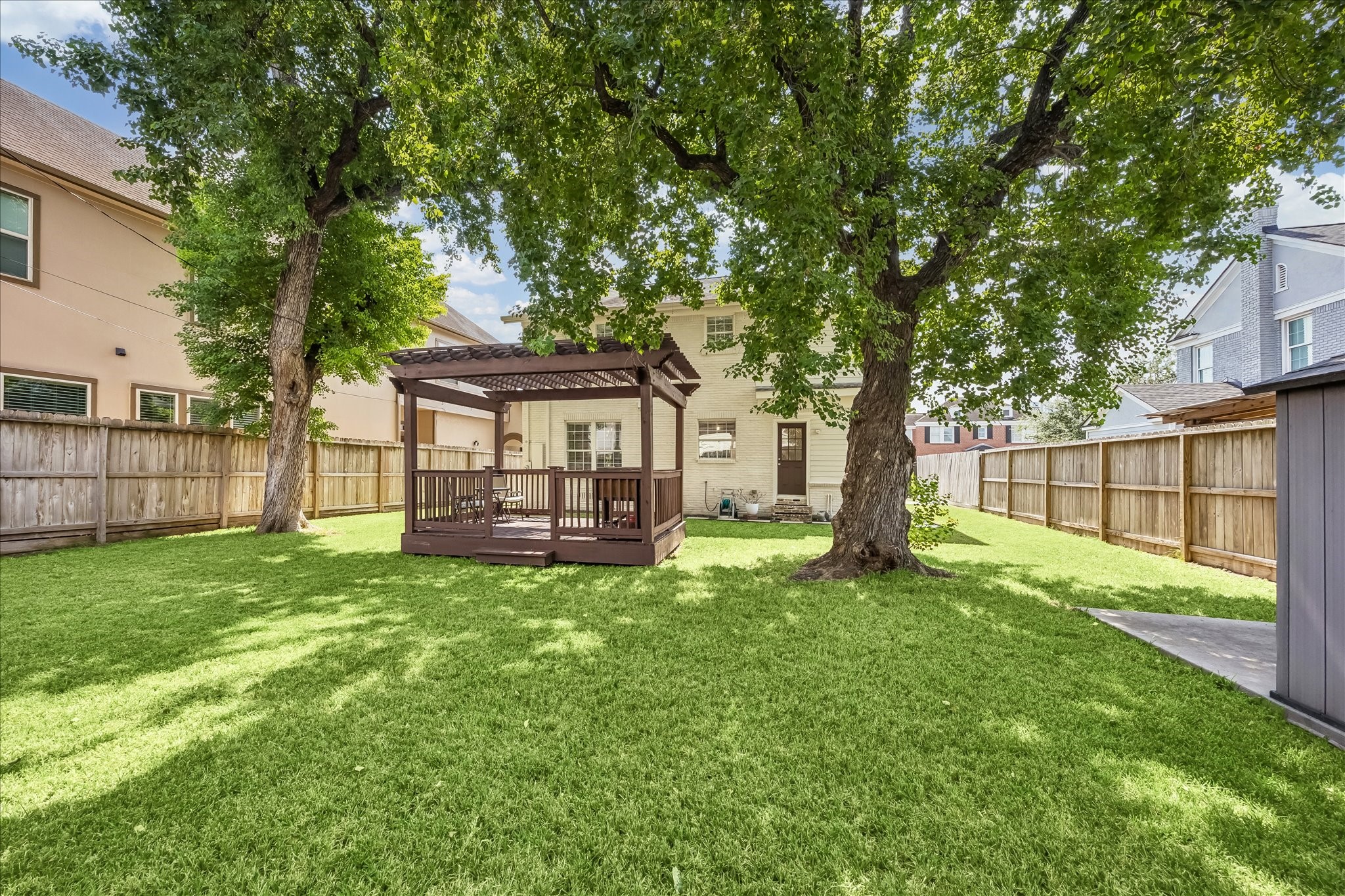Mature shade trees create a peaceful backdrop across the fenced yard, with the pergola positioned toward the rear for added separation.