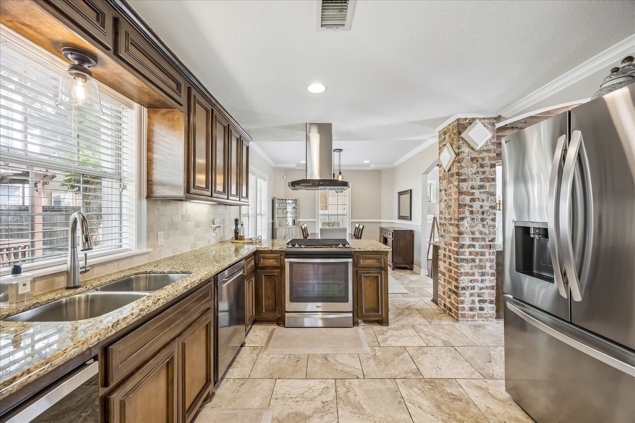 Granite countertops, rich wood cabinetry, stainless appliances, and gas cooking line the kitchen, with a brick column adding texture at the far end.