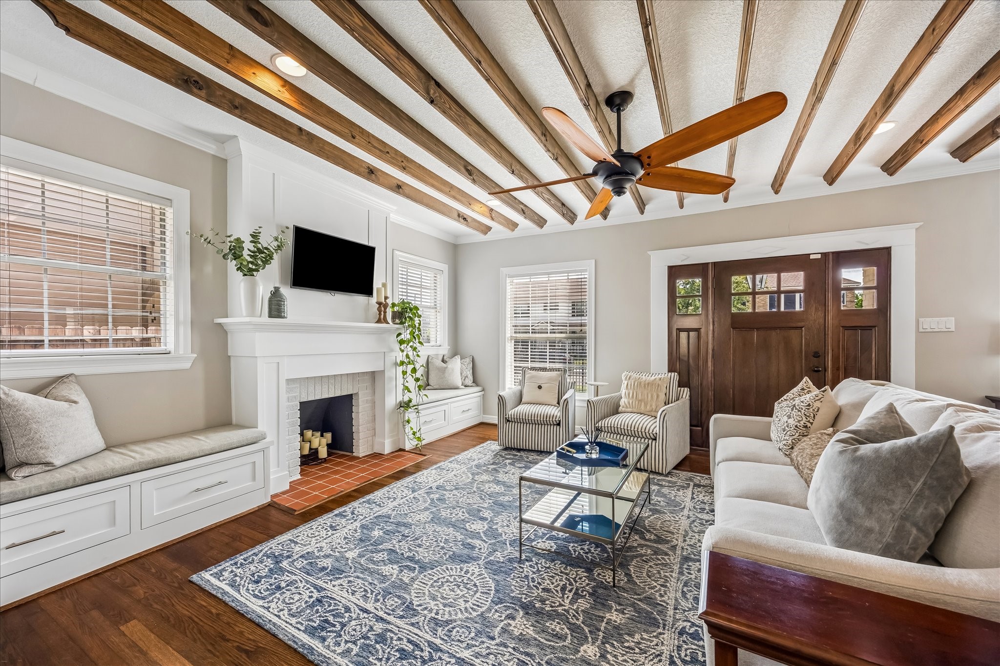 Natural light pours into the living room from multiple sides, highlighting the fireplace, detailed ceiling beams, and classic millwork.