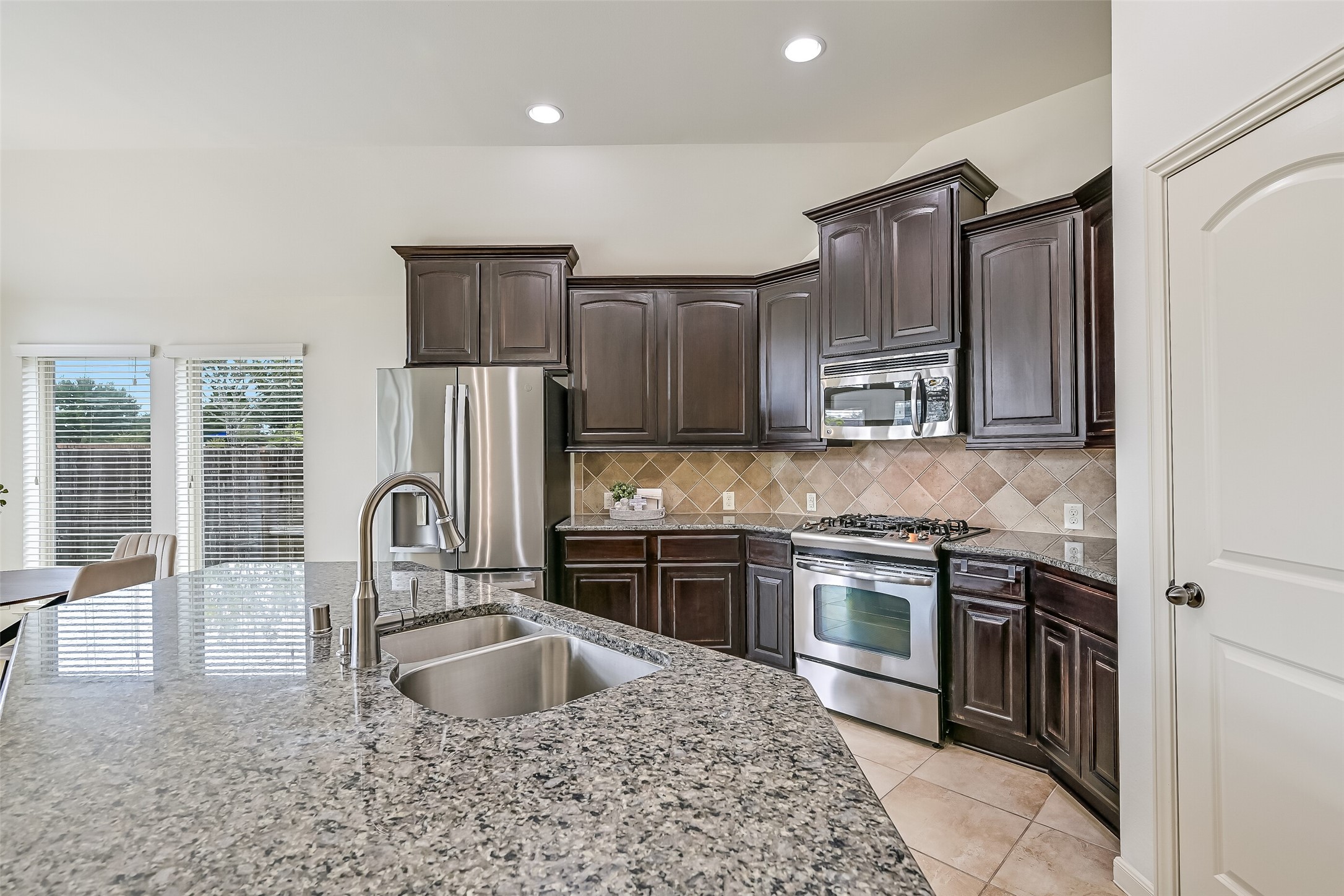 The kitchen features an abundance of dark-stained cabinetry with raised-panel doors, granite countertops & a decorative diagonal tile backsplash. Stainless steel appliances include a gas range, built-in microwave & French-door refrigerator. Recessed lighting brightens the workspace, with a pantry nearby.