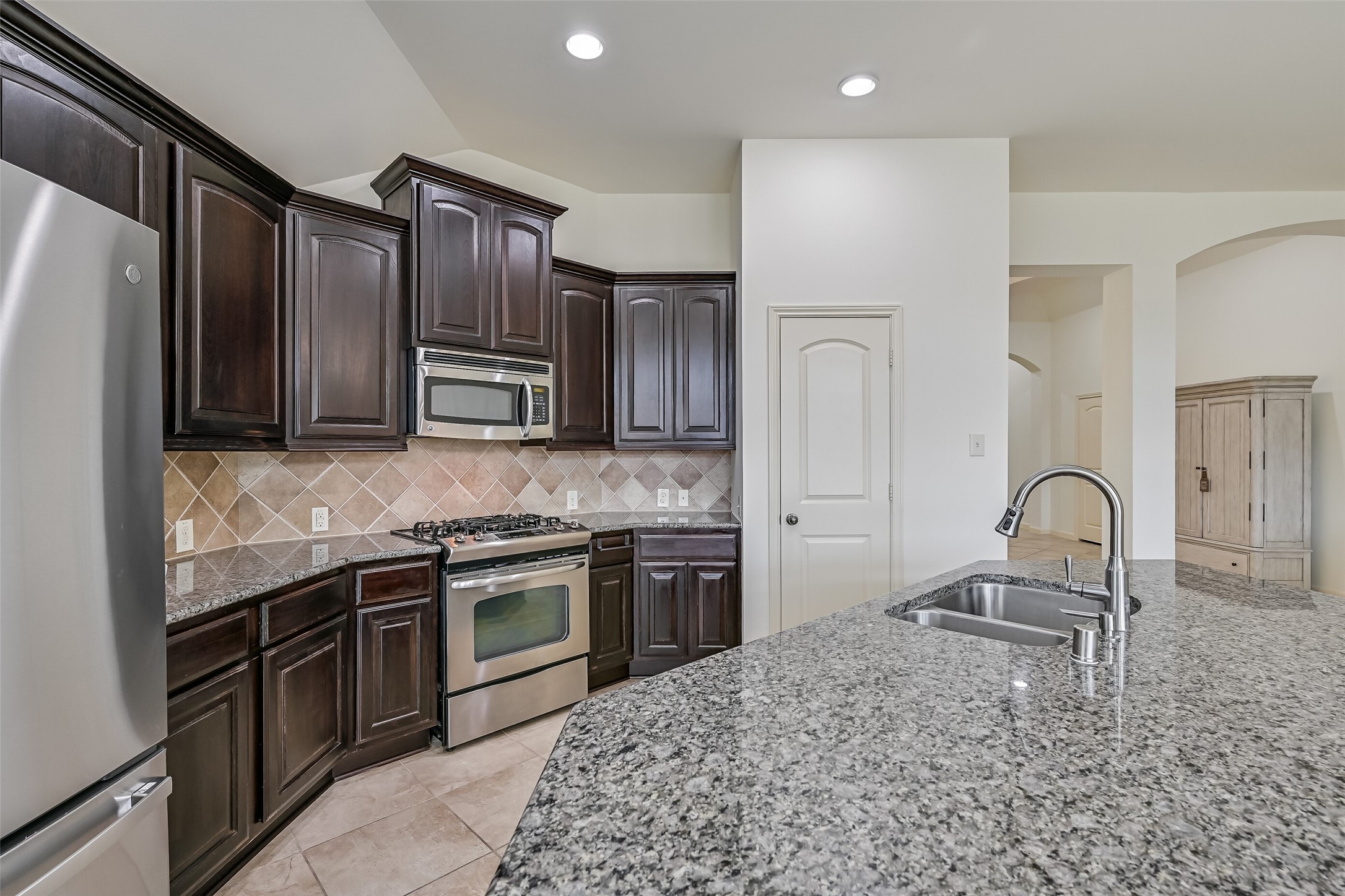 Beautiful granite countertops provide generous prep space throughout the kitchen, complemented by rich wood cabinetry & stainless steel appliances. The oversized island features a double-basin undermount sink with a high-arc faucet, while the gas range & decorative tile backsplash complete the cooking area.