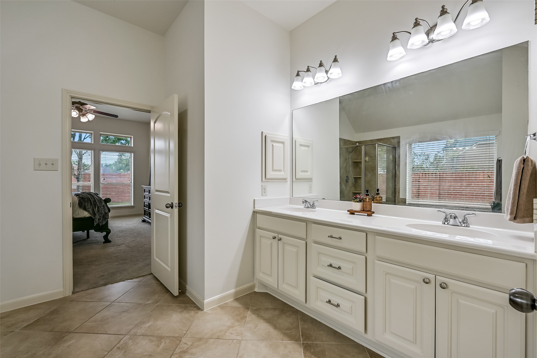 The primary bathroom features a long vanity with dual sinks, generous countertop space, white cabinetry & a large wall-to-wall mirror. Separate vanity lighting, neutral tile flooring & freshly painted walls create a bright, welcoming atmosphere, with convenient access to the adjoining primary bedroom.