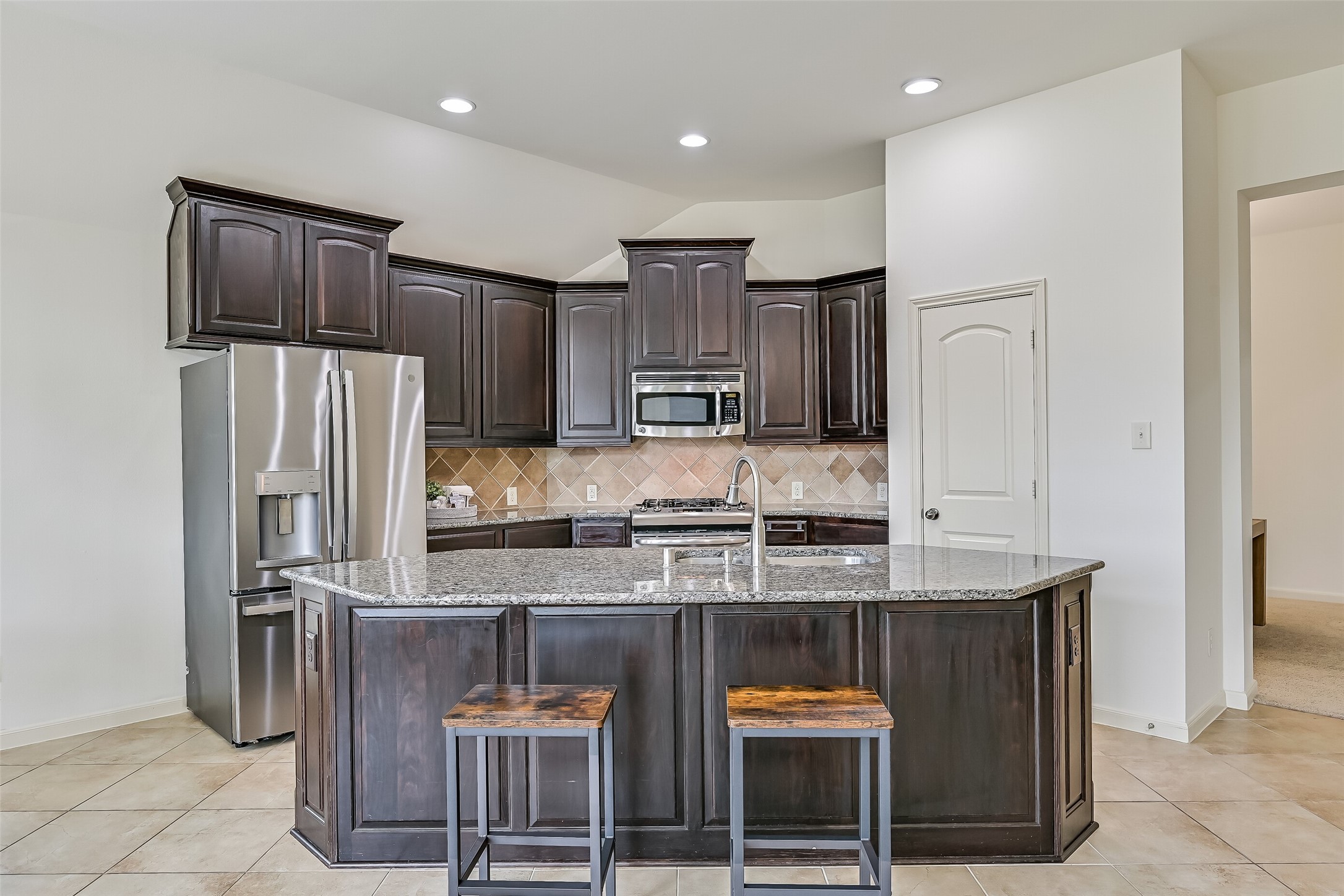 The spacious kitchen features an oversized granite island with breakfast bar seating, rich dark-stained cabinetry, stainless steel appliances, a gas range & decorative tile backsplash. Recessed lighting brightens the space, while the open design provides a seamless connection to the family room & breakfast area.