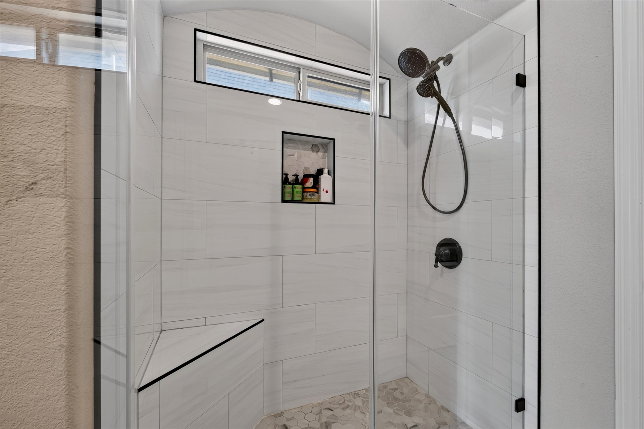 Enjoy the modern look of this glass-enclosed walk-in shower, featuring large-format wall tile, a decorative niche, and dark fixtures. A built-in bench adds convenience while the high window allows natural light to brighten the space.