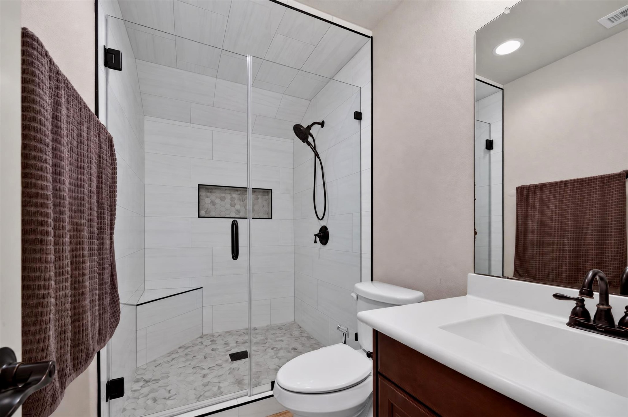 The secondary bathroom showcases a glass-enclosed walk-in shower with crisp white wall tile, decorative mosaic flooring, and dark fixtures. A white countertop and warm wood cabinetry complete the clean, contemporary look.
