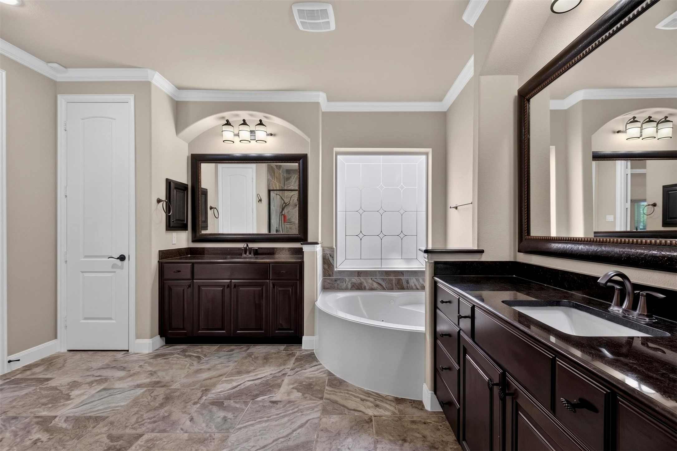 This alternate view highlights the bathroom's thoughtful arrangement, with dual vanities positioned on opposite sides of the room and the soaking tub nestled between them. Coordinating cabinetry, framed mirrors, and decorative tile create a cohesive appearance throughout.