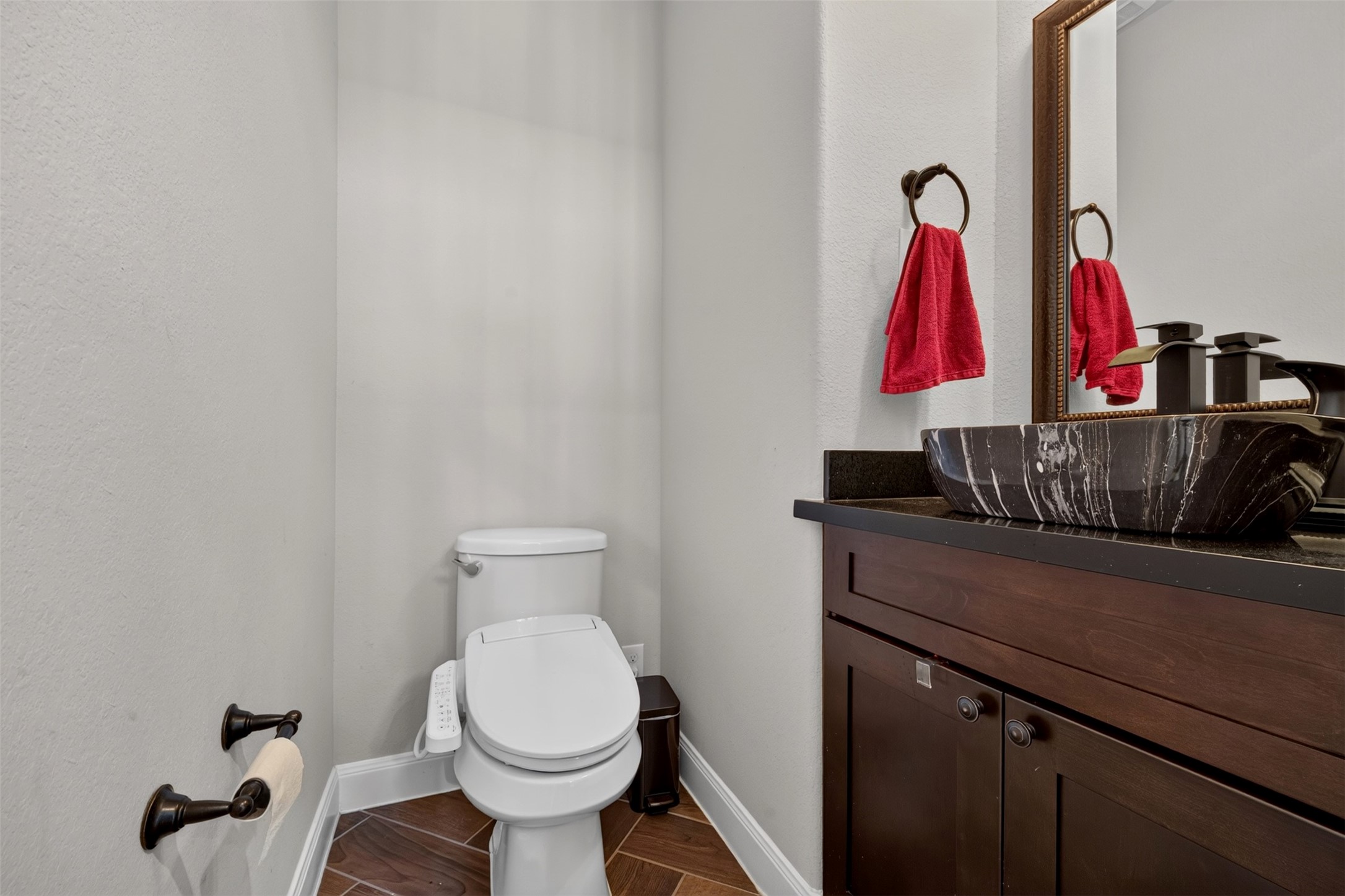 A stylish vessel sink rests atop a dark countertop, paired with a rich wood vanity and a framed mirror. Warm wood-look flooring and coordinating fixtures give this conveniently located half bath a distinctive finish.
