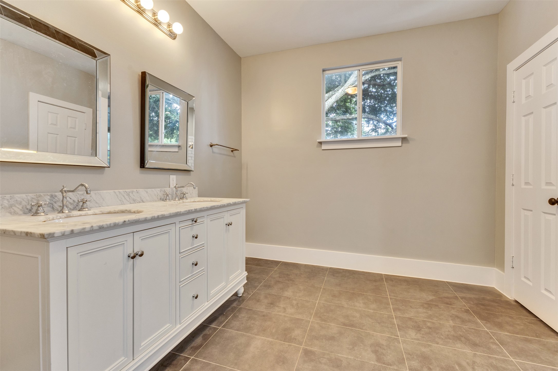 Spacious bathroom featuring a dual-sink vanity with elegant marble countertops, modern lighting, and a large mirror, complemented by neutral walls and stylish tile flooring.