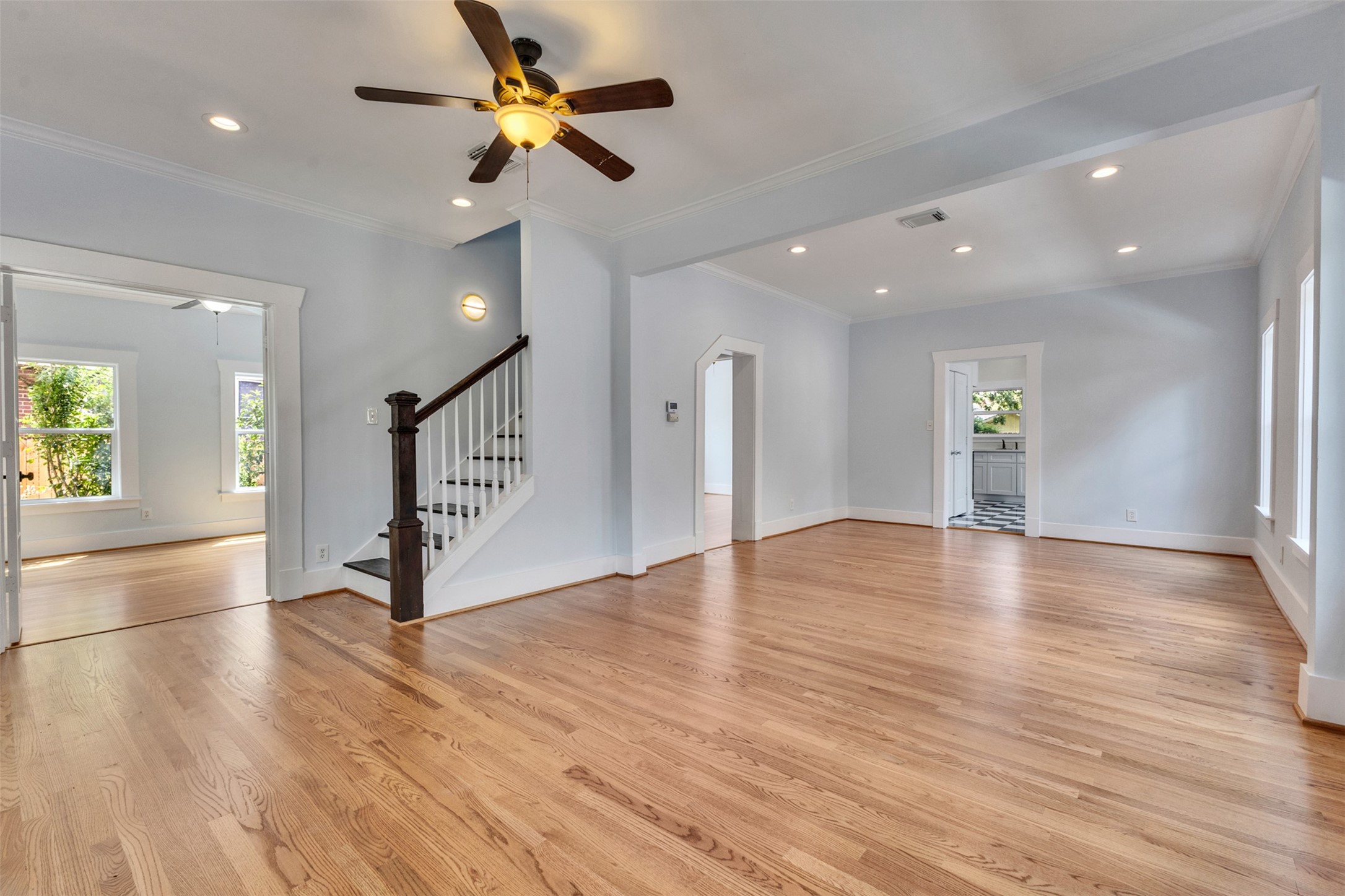 Spacious open-concept living area featuring hardwood floors, a ceiling fan, and abundant natural light, with easy access to adjacent rooms and a staircase leading upstairs.