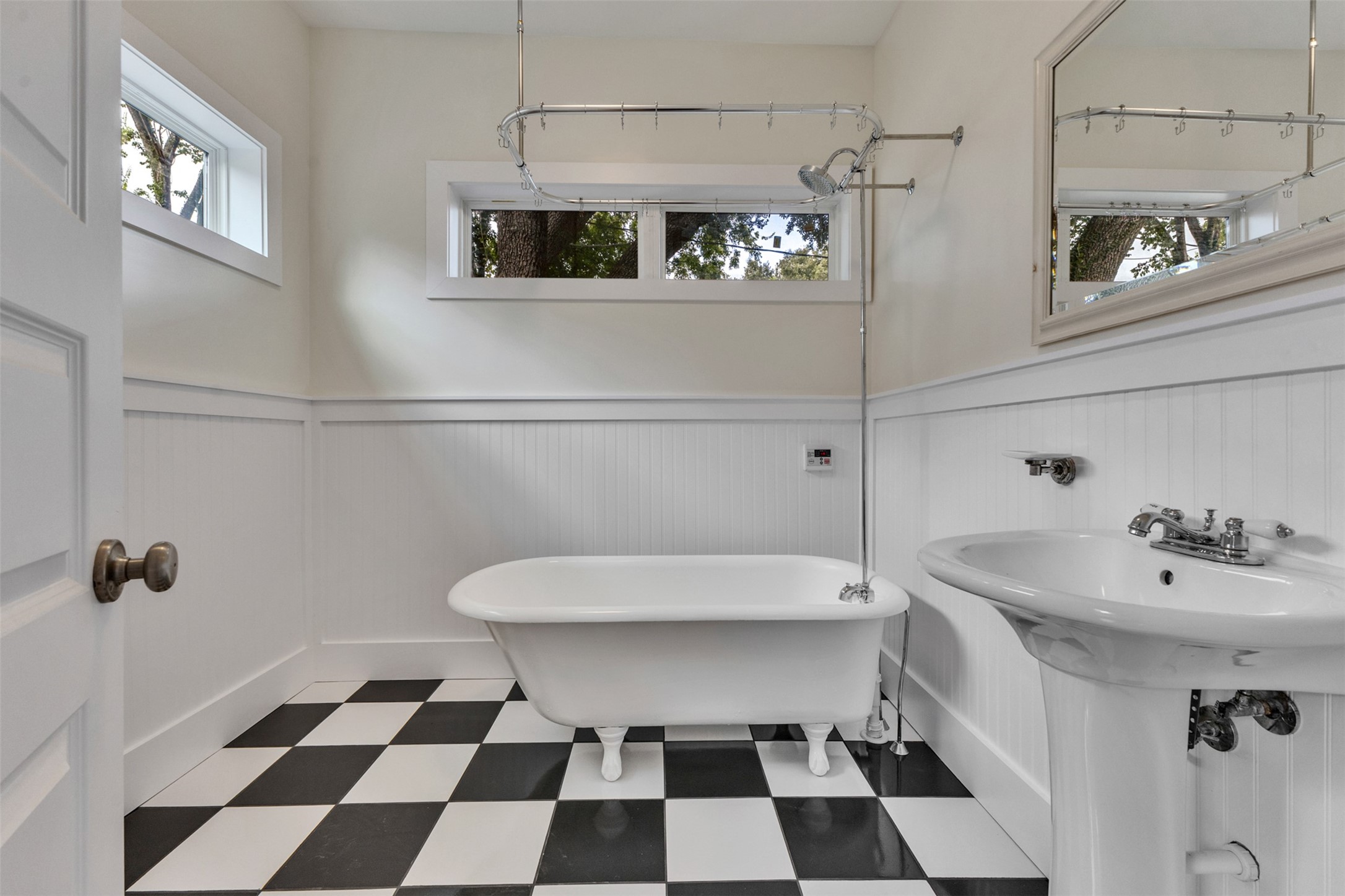 Charming bathroom featuring a classic clawfoot tub, elegant pedestal sink, and stylish black and white checkered flooring, complemented by natural light through large windows.