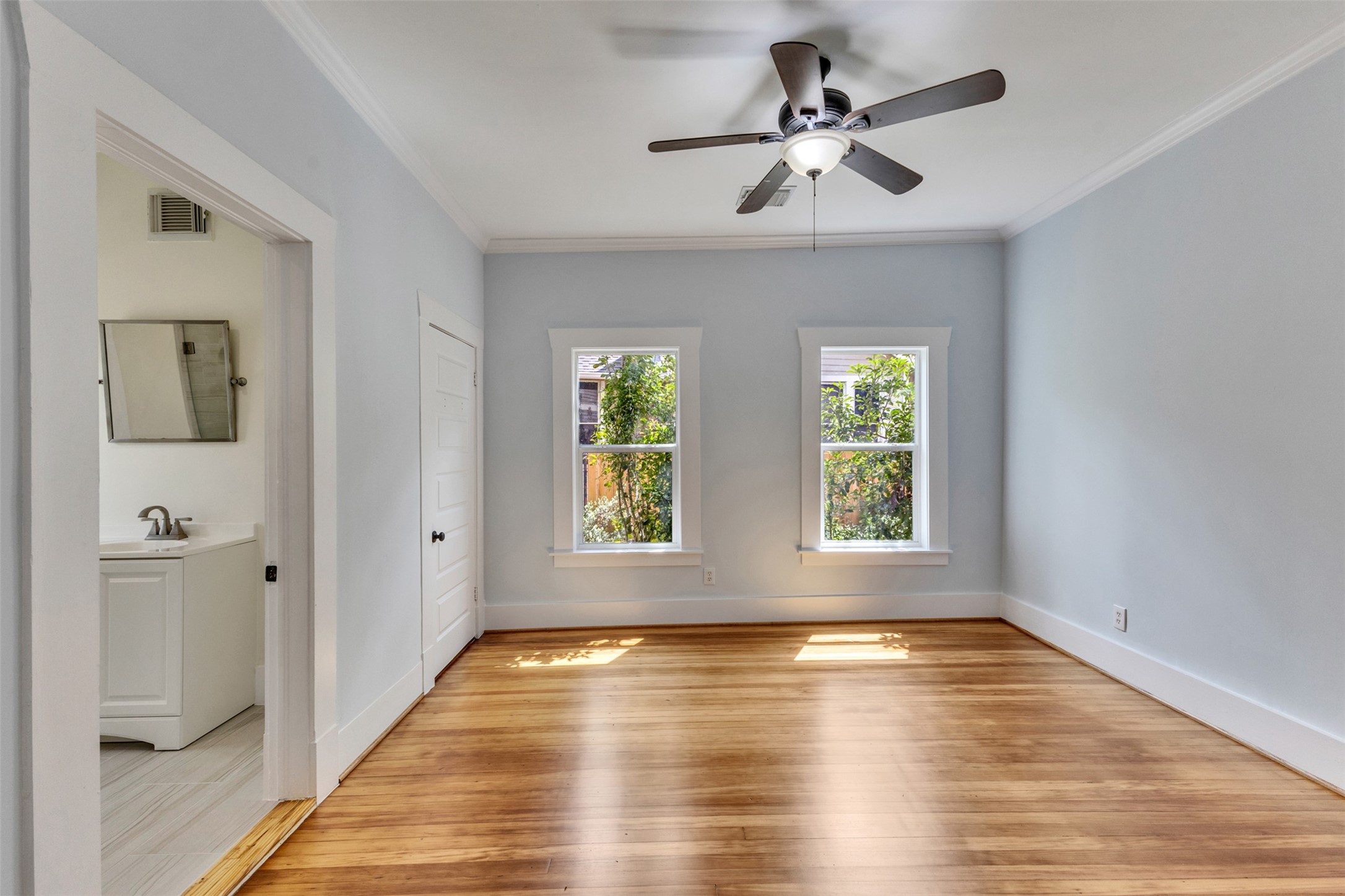 Bright and airy room featuring hardwood floors, large windows for natural light, and a ceiling fan, with an adjoining bathroom for convenience.