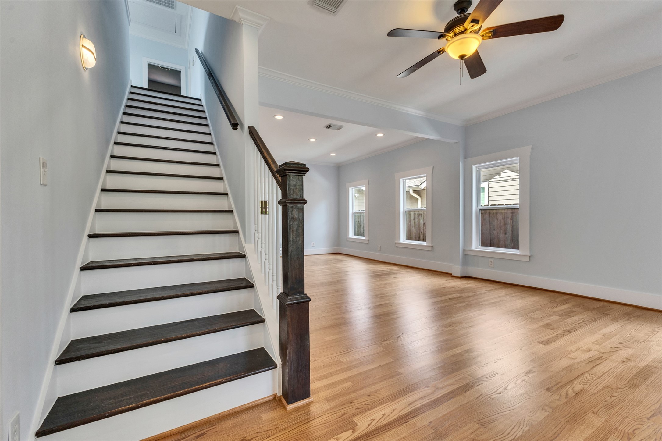 Spacious living area with gleaming hardwood floors and a ceiling fan, complemented by natural light from multiple windows. A classic staircase with dark wood accents adds elegance and leads to the upper level.