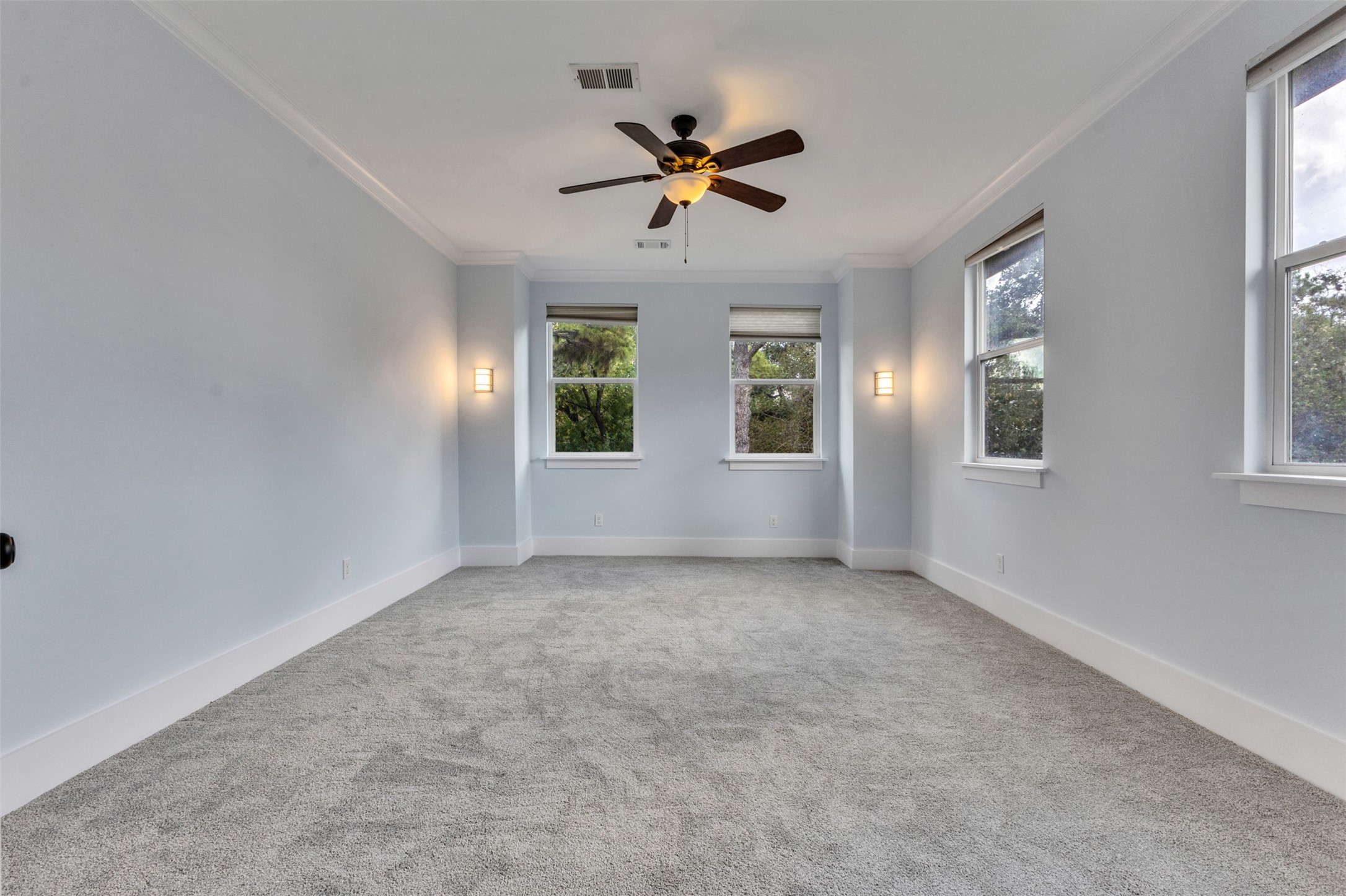 Spacious room with soft gray carpeting and ample natural light from multiple windows, featuring elegant wall sconces and a ceiling fan.