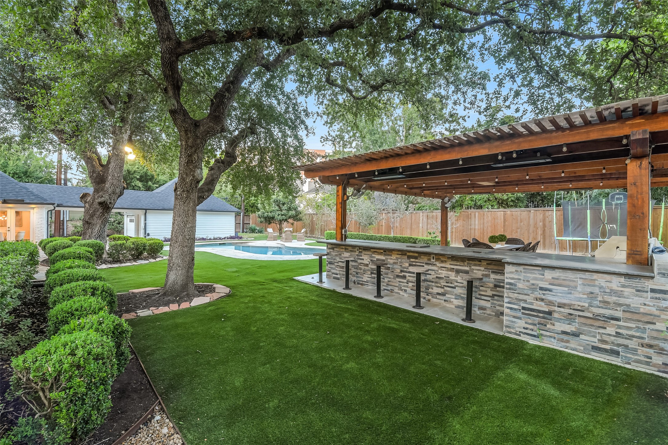 Another view of the beautifully maintained backyard showcasing lush landscaping, mature shade trees, a sparkling pool, and an expansive covered outdoor kitchen with bar seating.