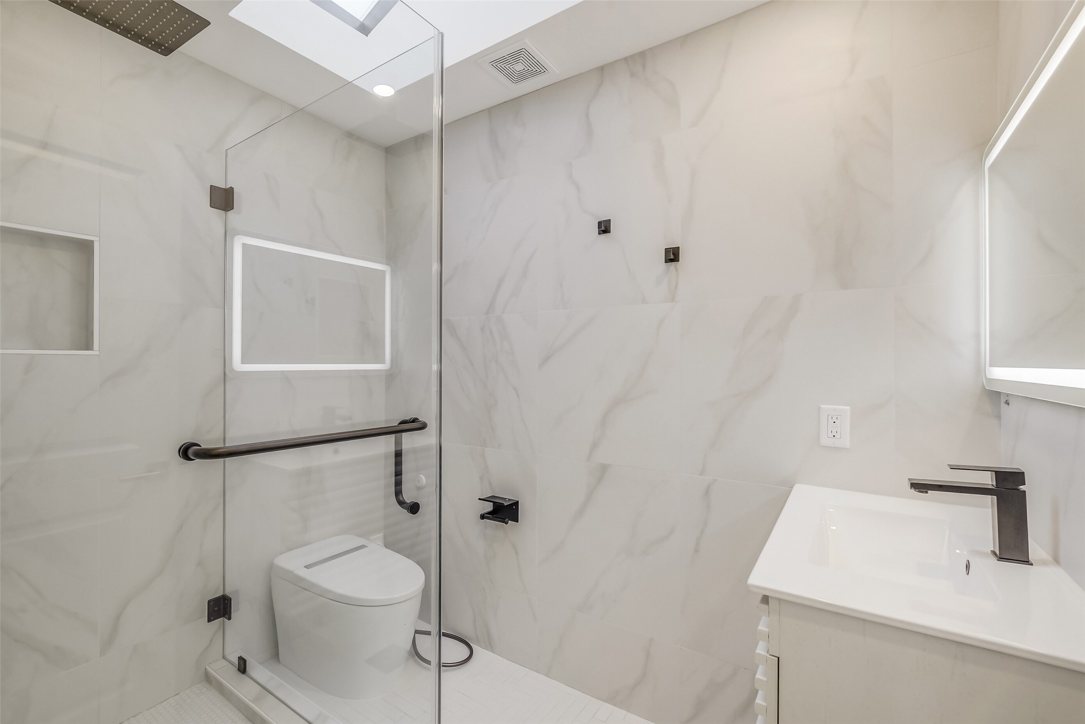 Contemporary secondary bathroom en-suite featuring marble-look tile, a glass-enclosed shower with rainfall showerhead, sleek fixtures, and a modern vanity with illuminated mirror.