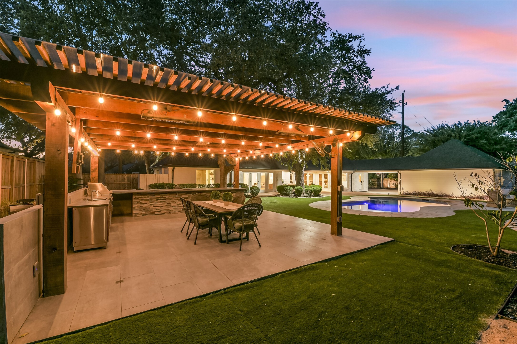 Designed for year-round entertaining, this impressive outdoor kitchen and dining area features a wood pergola, warm string lighting, stainless steel appliances, and picturesque views of the illuminated pool.