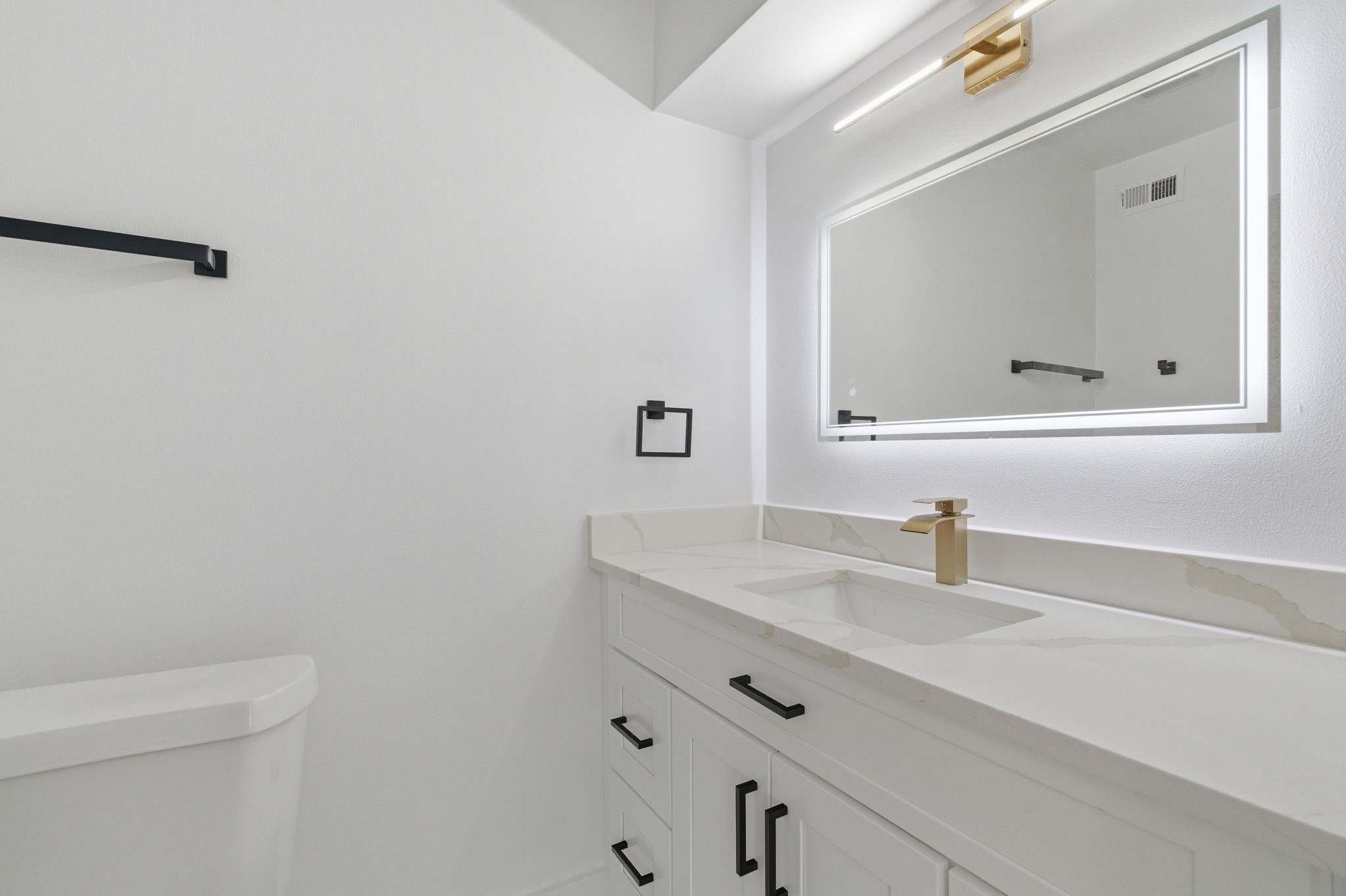 The secondary bathroom carries the same updated style with an illuminated mirror, gold tone faucet, and black hardware.