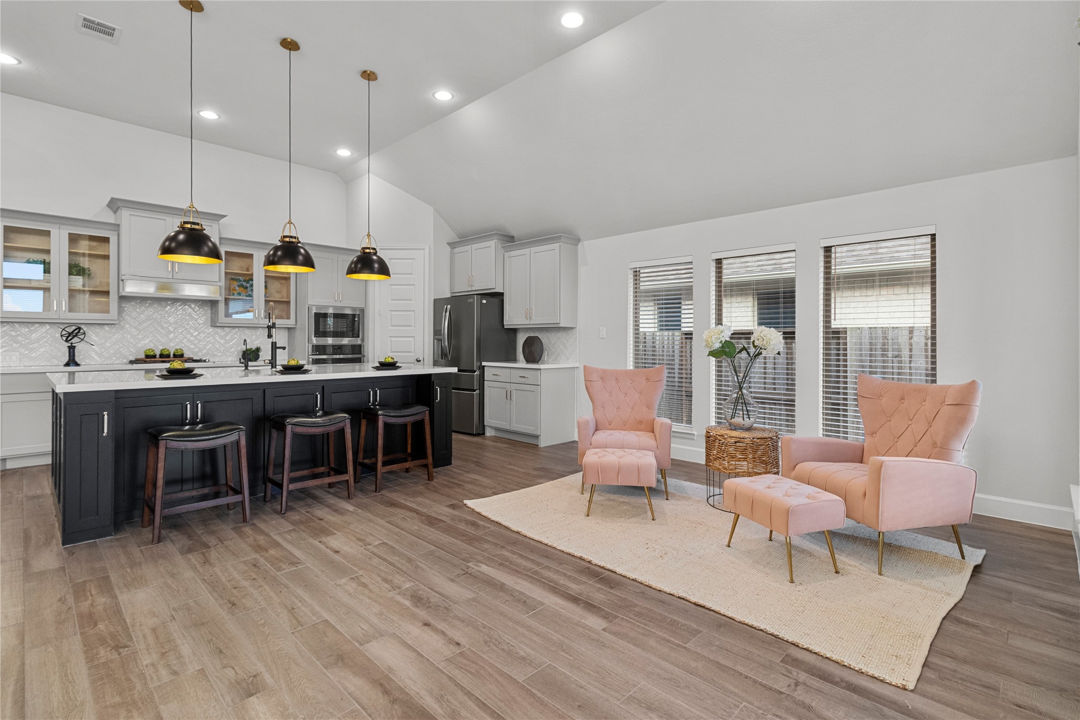 A modern open-concept living area features a sleek kitchen with a large island, pendant lighting, and ample seating. The space seamlessly flows into a cozy sitting area with plush pink chairs and abundant natural light.