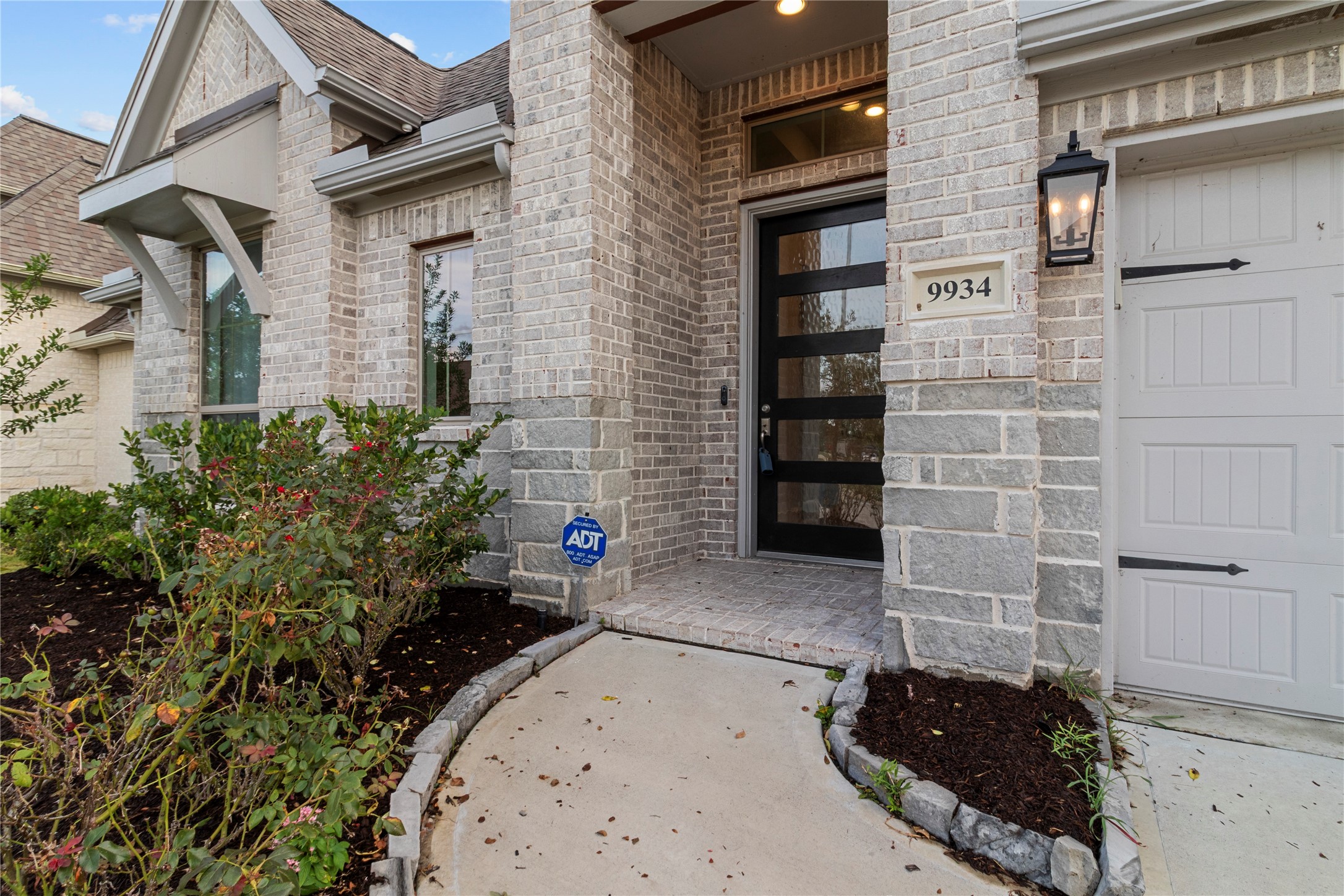 Charming entrance with a modern glass door, elegant brickwork, and a welcoming porch, complemented by tasteful landscaping and a classic lantern-style light fixture.
