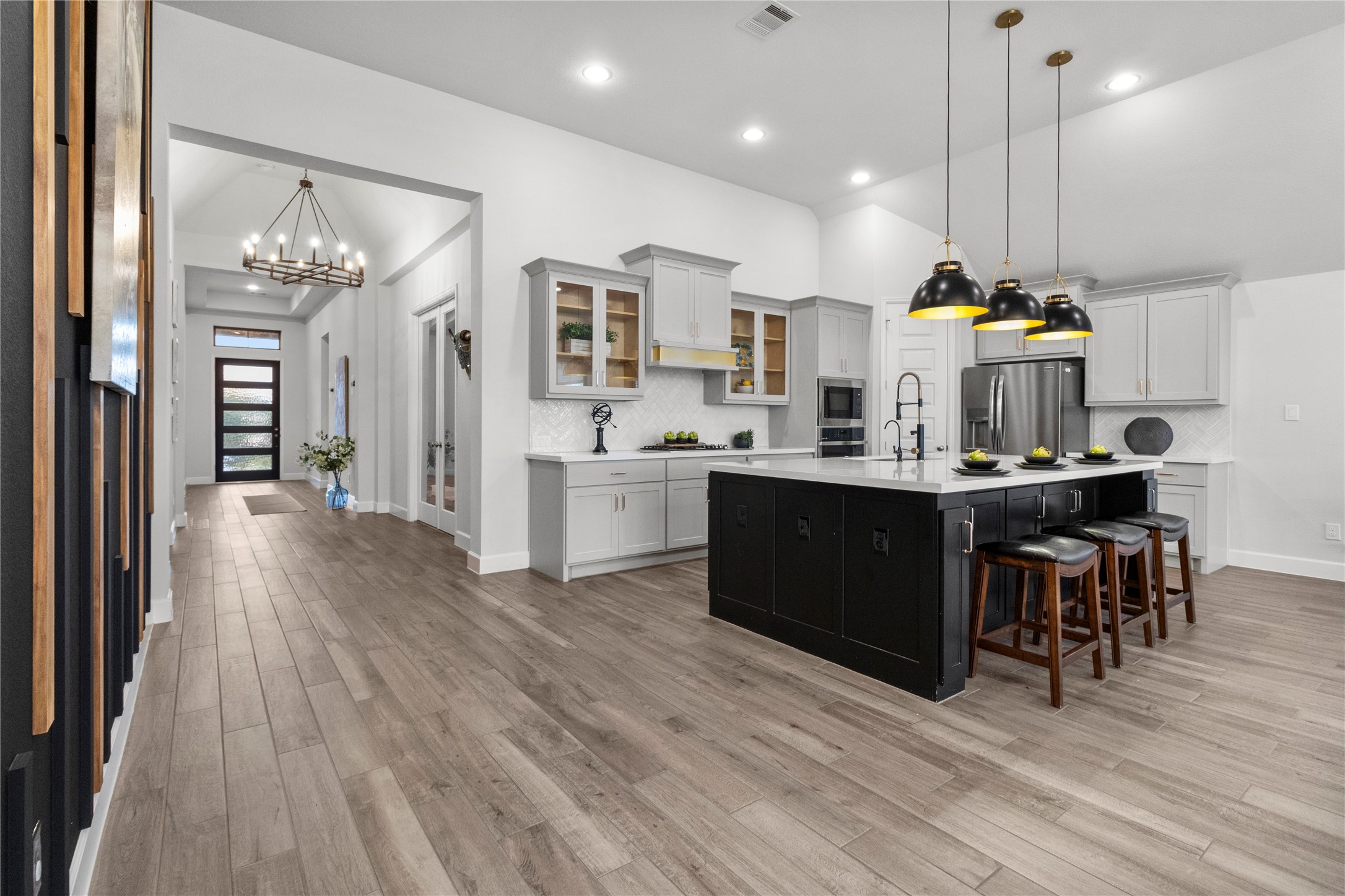 Spacious modern kitchen with sleek cabinetry, a large island with seating, and elegant pendant lighting, seamlessly flowing into a welcoming hallway adorned with stylish chandeliers and abundant natural light.