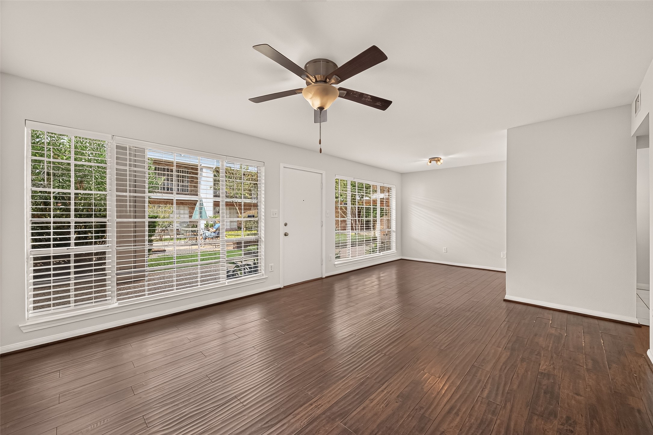 Spacious living area with rich hardwood floors, abundant natural light from large windows, and a modern ceiling fan, offering a warm and inviting atmosphere.