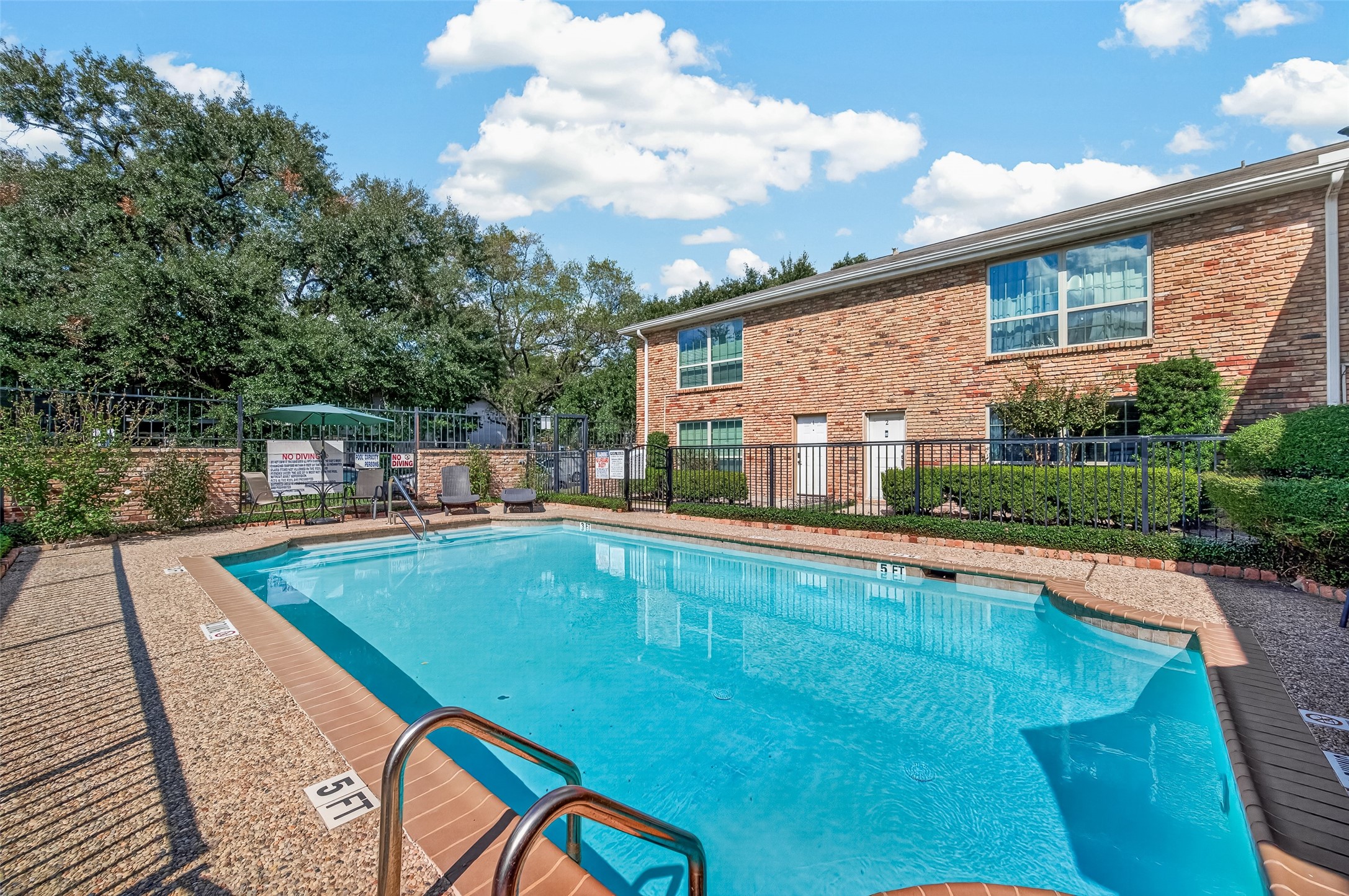 Enjoy a refreshing swim in this inviting community pool, nestled beside a charming brick building and surrounded by lush greenery.