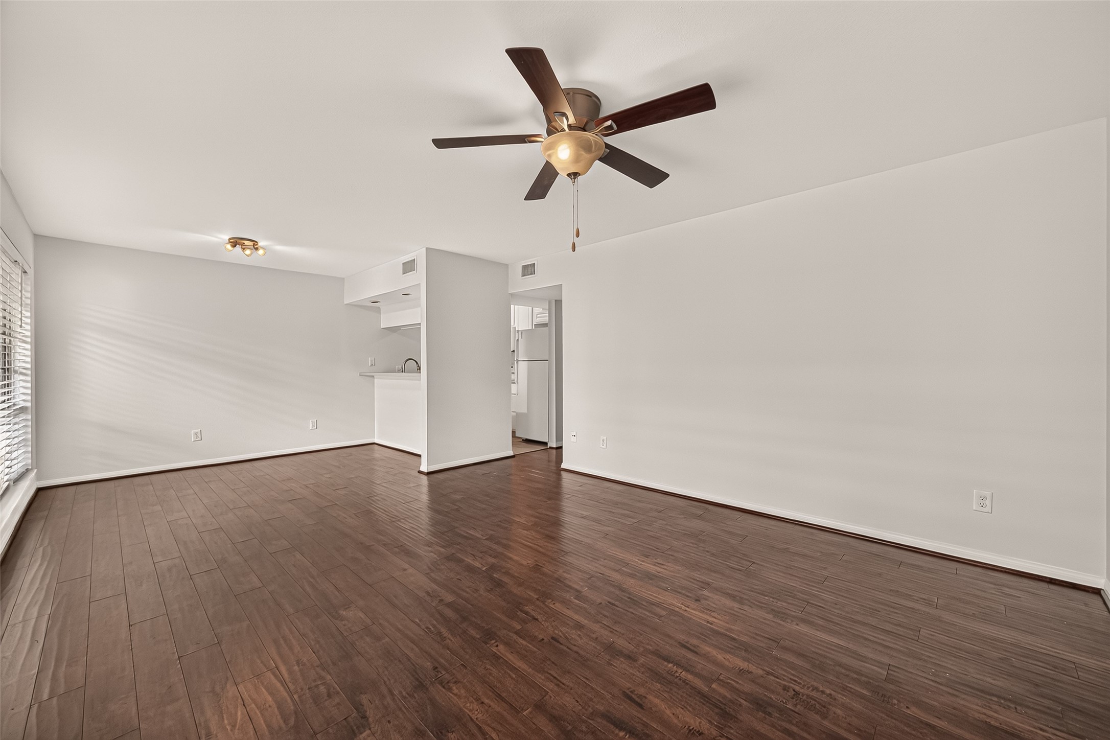 Spacious living area with elegant dark wood flooring and a ceiling fan, seamlessly flowing into a cozy kitchen nook.