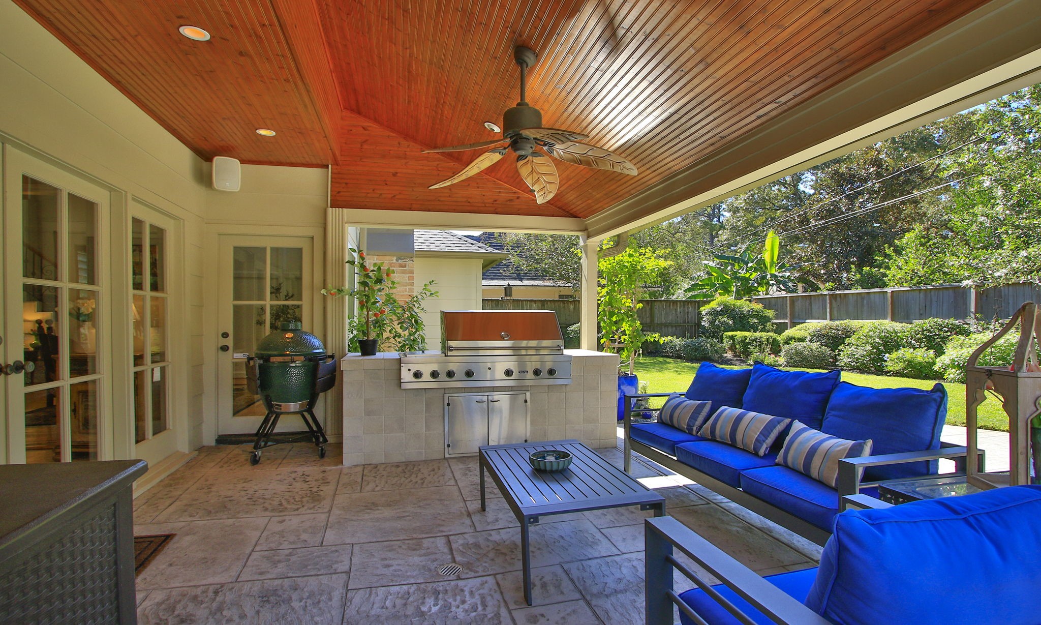 Experience outdoor living at its finest with this beautifully designed patio featuring a built-in grill, and a ceiling fan under a warm wood-paneled ceiling, all set against a lush garden backdrop.
