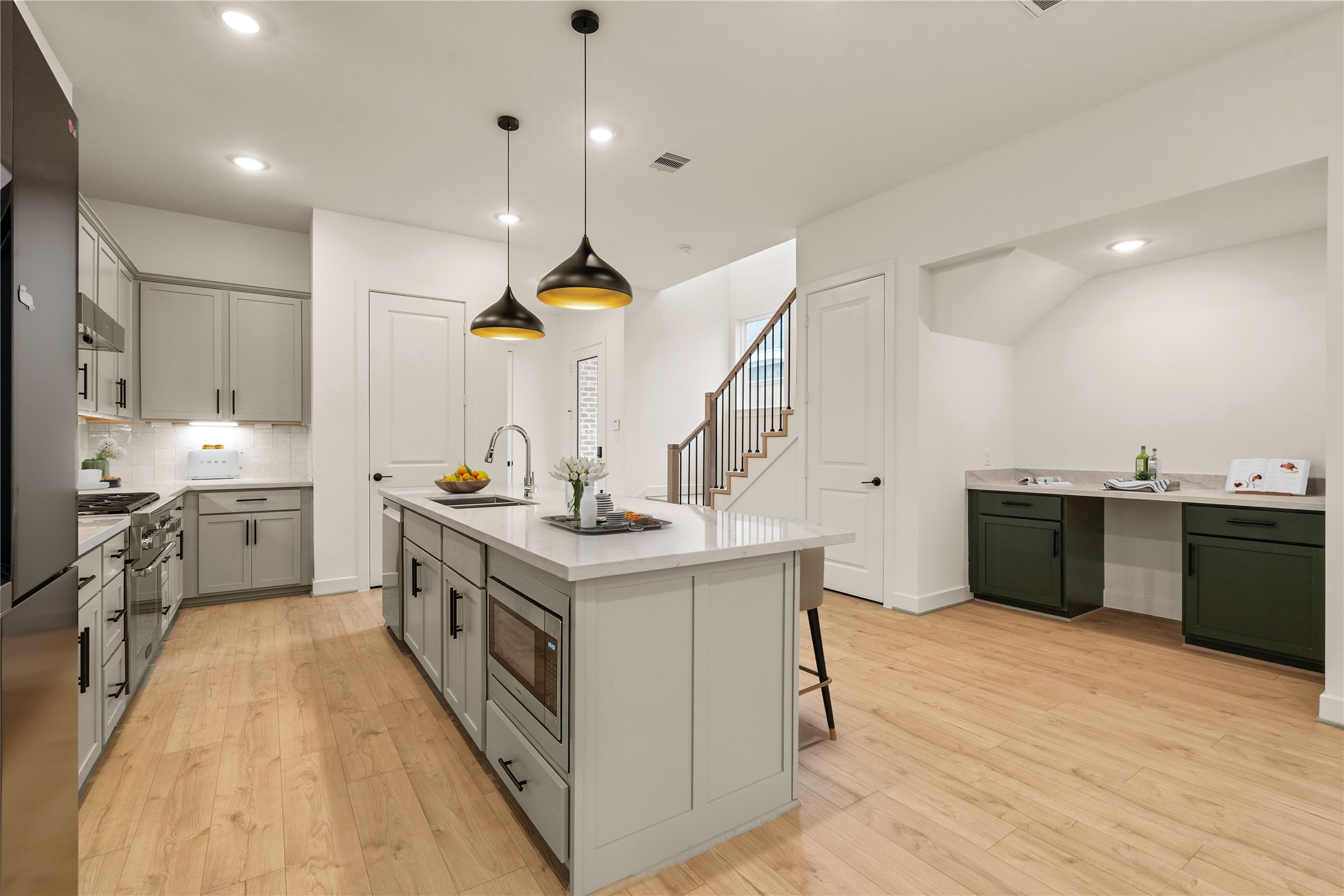Gourmet kitchen and bar area  spacious quartz-topped island with under-cabinet microwave, matte black pendant lights, soft-grey shaker cabinetry, and an adjacent wet bar nook set over light oak flooring.