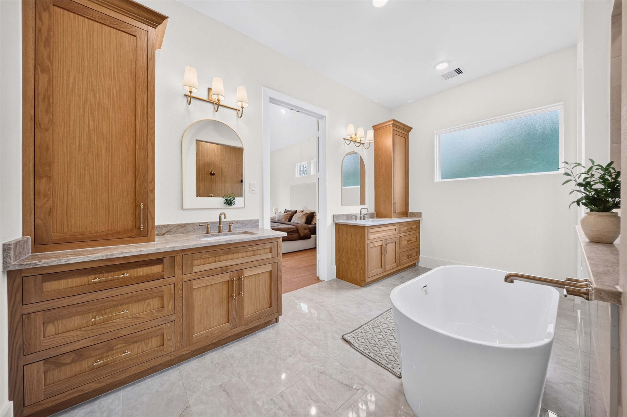 The primary bath centers on a freestanding tub between dual wood vanities, with pale tile and a wide window bringing in natural light.