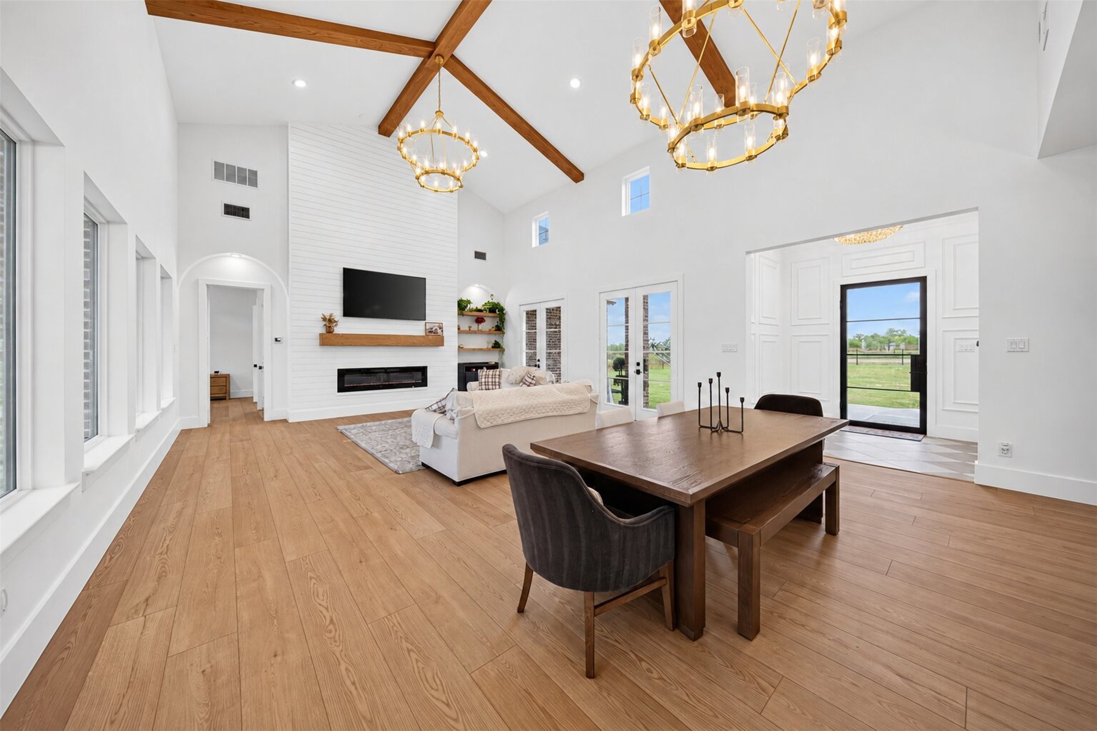 Tall windows frame yard views across the open living and dining areas, unified by a vaulted ceiling and exposed wood beams.
