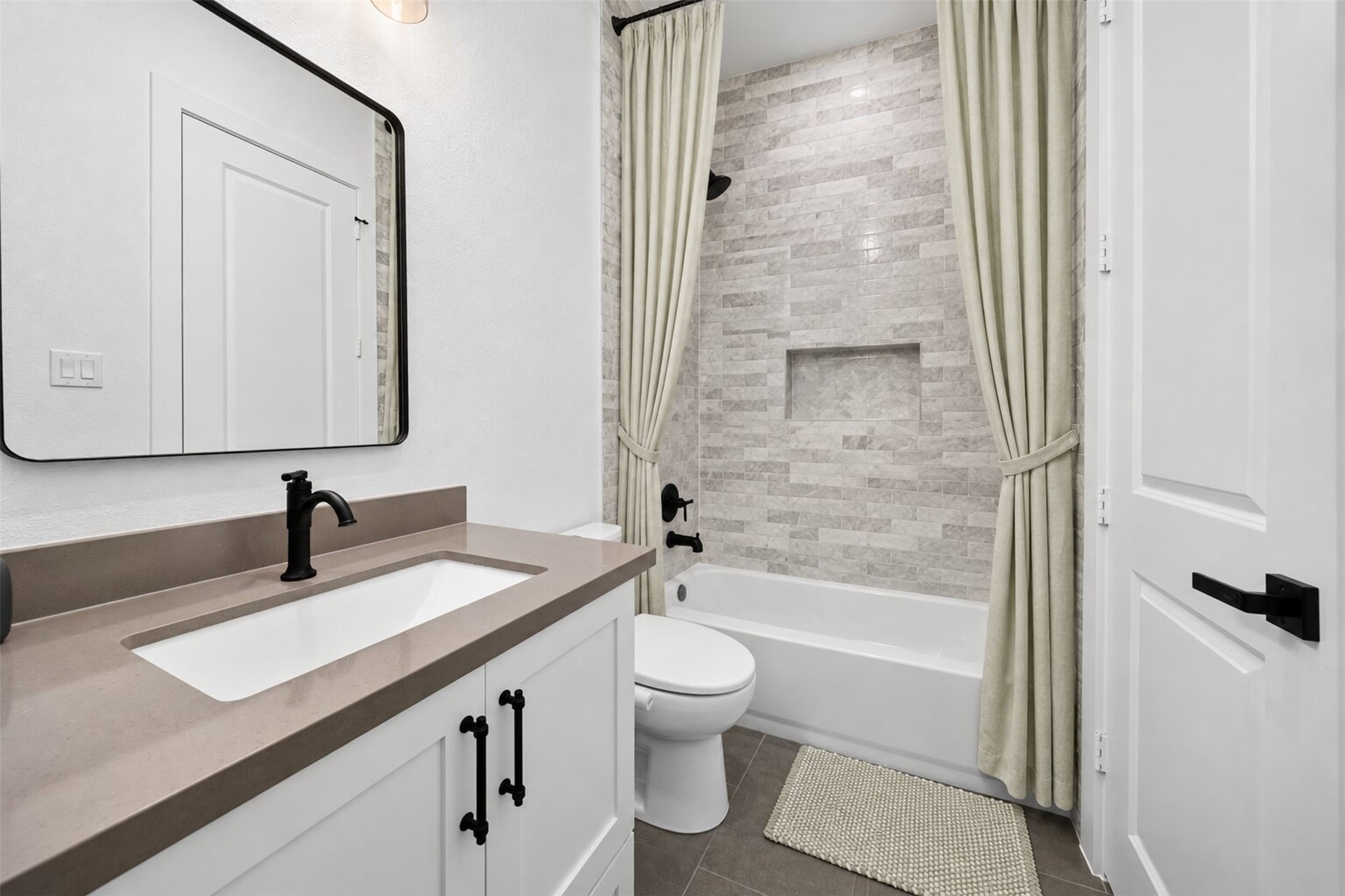 Light marble-look tile wraps the tub and shower, paired with a pale vanity and dark fixtures for a clean, modern finish.