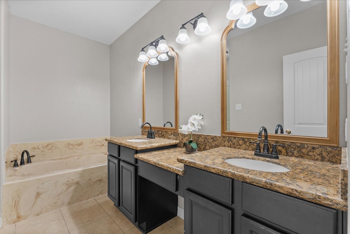 The primary bath features a double vanity with granite counters and two framed mirrors under wall-mounted light fixtures, plus a separate soaking tub for a spa-like retreat. Dark cabinetry and tile flooring round out the finishes.