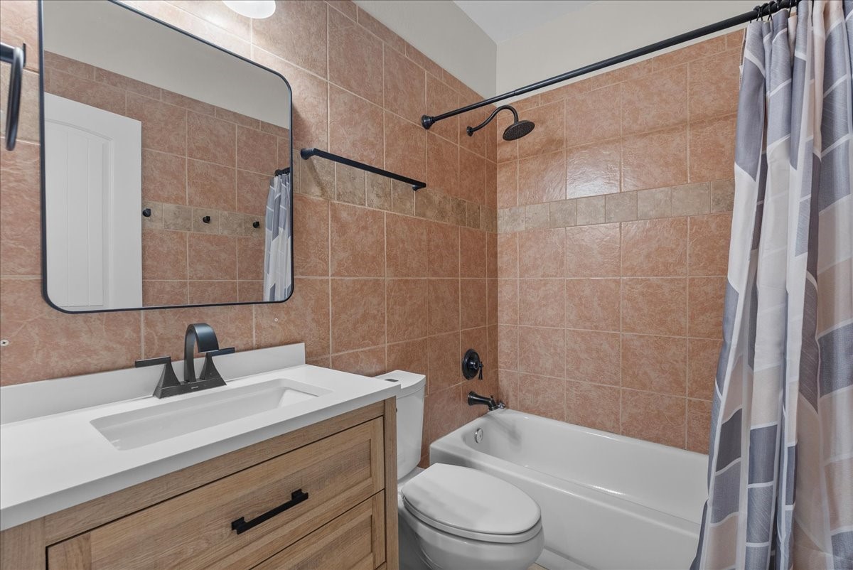 A full tub/shower combo is finished with floor-to-ceiling tile surround and a built-in towel bar, paired with a wood vanity topped in quartz and an updated matte-black faucet. A large framed mirror and overhead lighting complete the space.