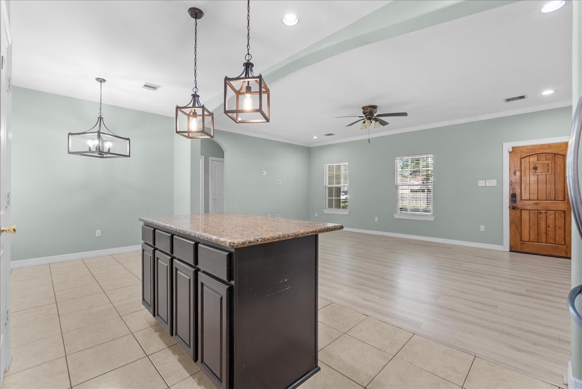 Three pendant fixtures illuminate the granite island, which opens directly to the vaulted living area beyond. Wide-plank flooring picks up where the kitchen tile leaves off, paired windows bring in natural light, and a ceiling fan plus the wood front entry door anchor the far end of the room.