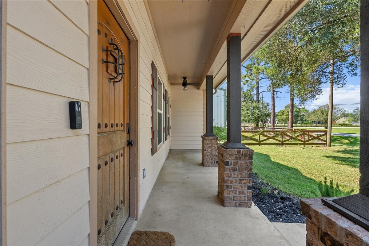 The arched wood door with wrought-iron hardware and a video doorbell sets a secure, custom tone right at the threshold, while the long covered walkway connects seamlessly to the porch beyond, framing clean sightlines out to the fenced yard and treeline.